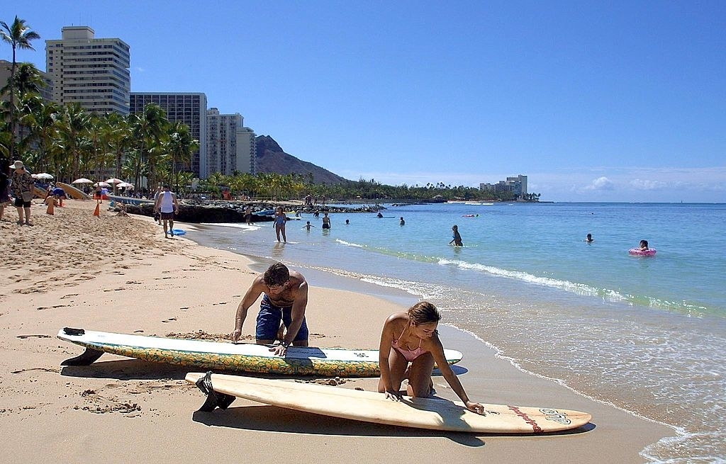 People prepare surfboards on a busy beach with hotels and palm trees in the background. The shoreline is dotted with beachgoers enjoying the sunny day