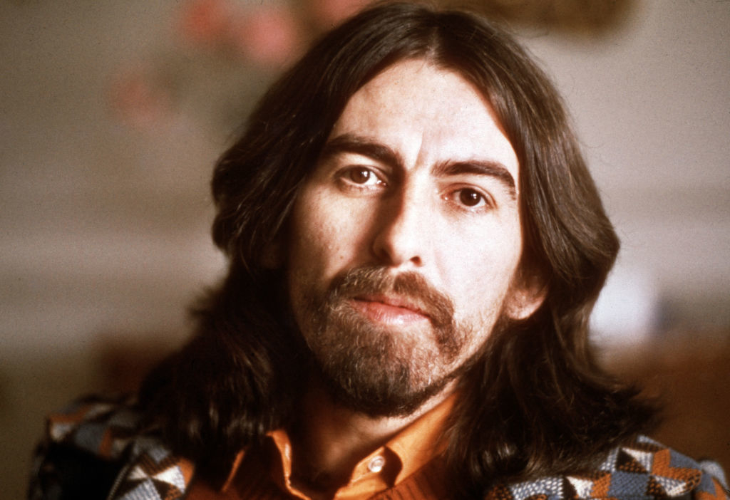 George Harrison with long hair and a beard, wearing a patterned jacket over a collared shirt