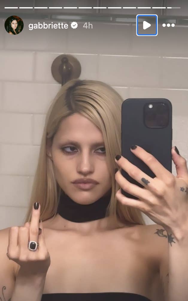 Gabriette in a strapless outfit takes a selfie in a bathroom, showing her ring finger to the camera