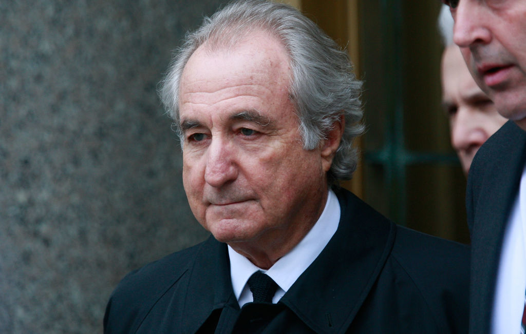 Bernie Madoff in a dark coat and tie, looking serious with another unidentified person partially visible in the background