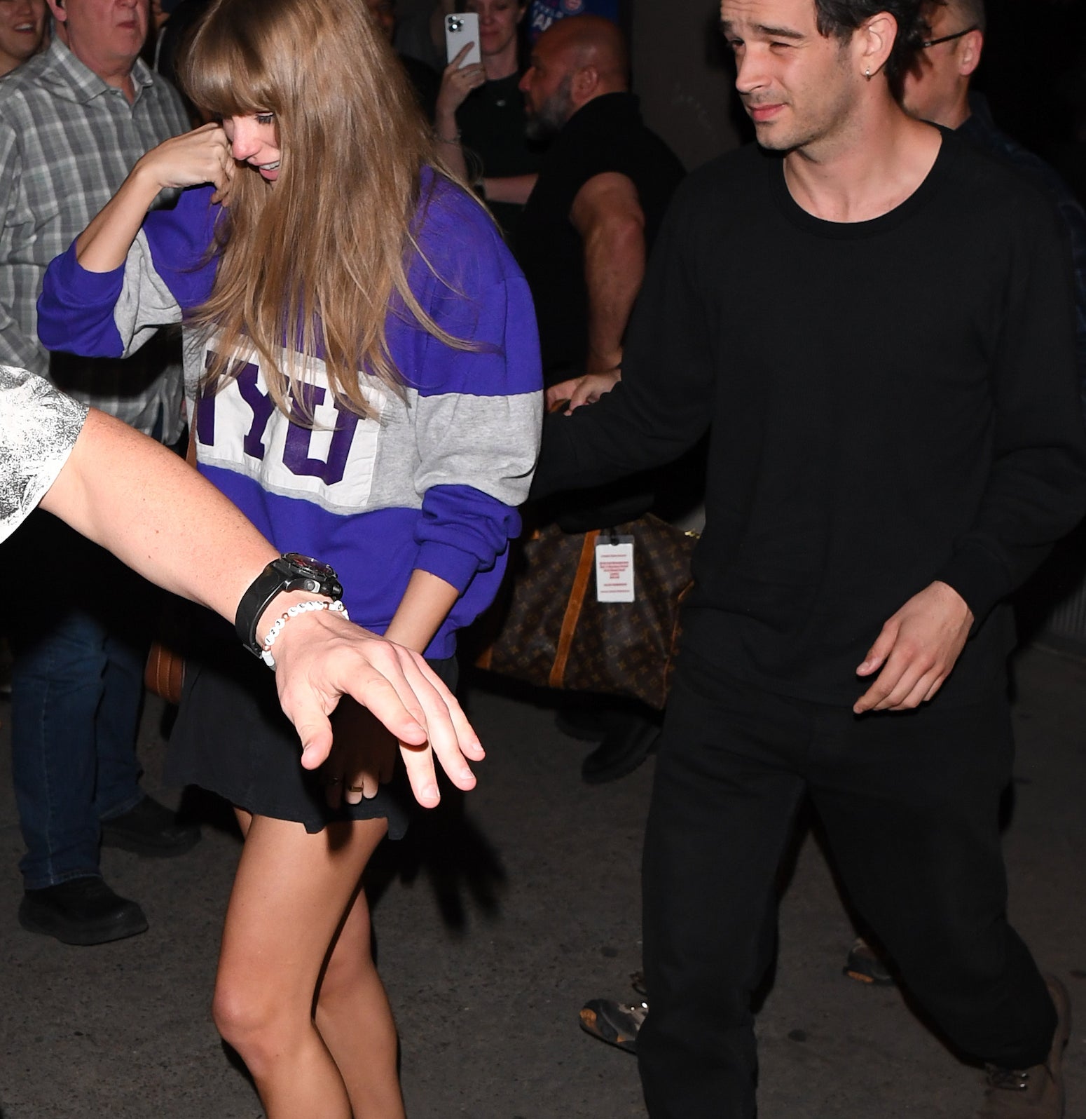 Taylor Swift is walking and covering her face with her hand. She is wearing a casual sweater with &quot;NYU&quot; on it and a skirt. She&#x27;s holding hands with Matty Healy, who is in a black outfit. They are both surrounded by fans and photographers