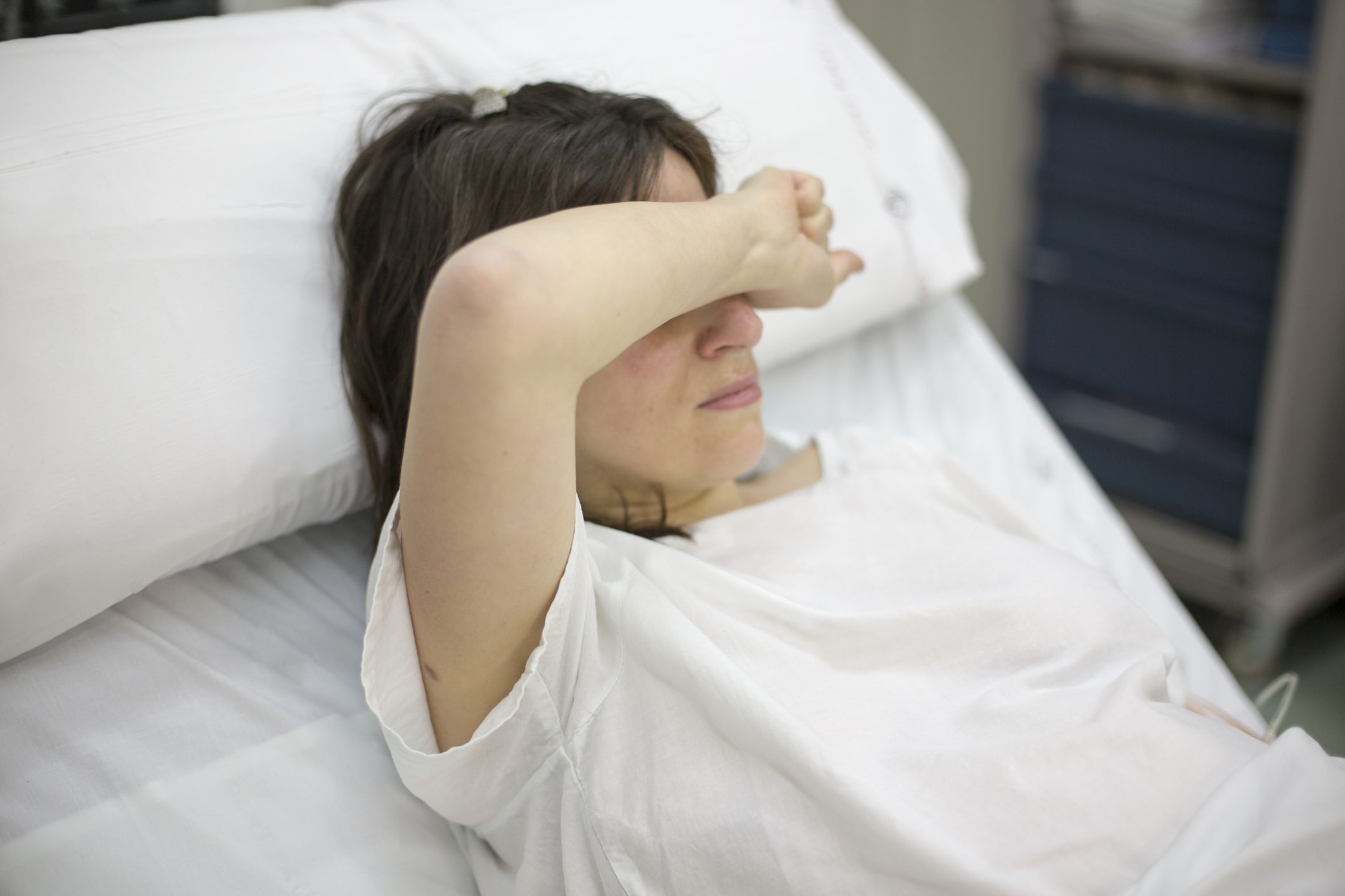 A person lies in a hospital bed with their arm covering their eyes, appearing to shield from light or rest