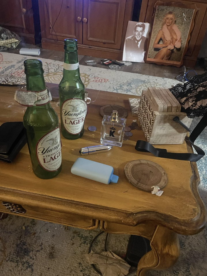 A cluttered coffee table with two beer bottles, a blue bottle, a coaster, coins, a perfume bottle, and various scattered items. Marilyn Monroe photo in the background