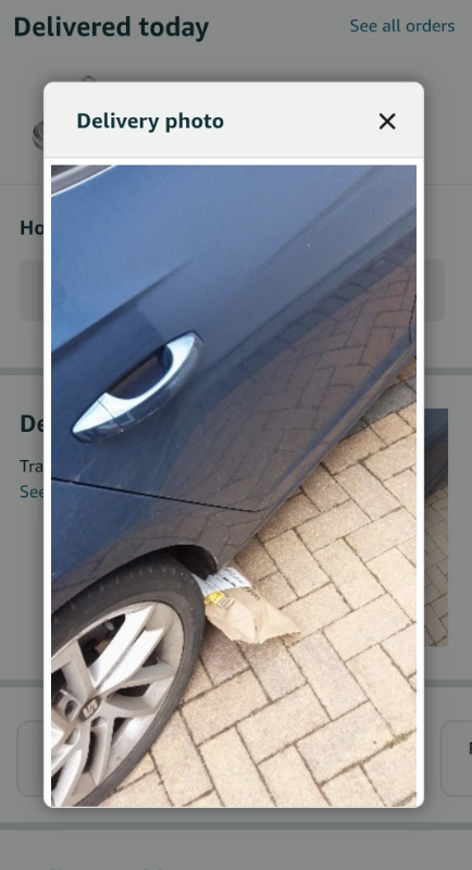Image shows a close-up of a blue car parked on a brick driveway with a bag placed near the rear wheel