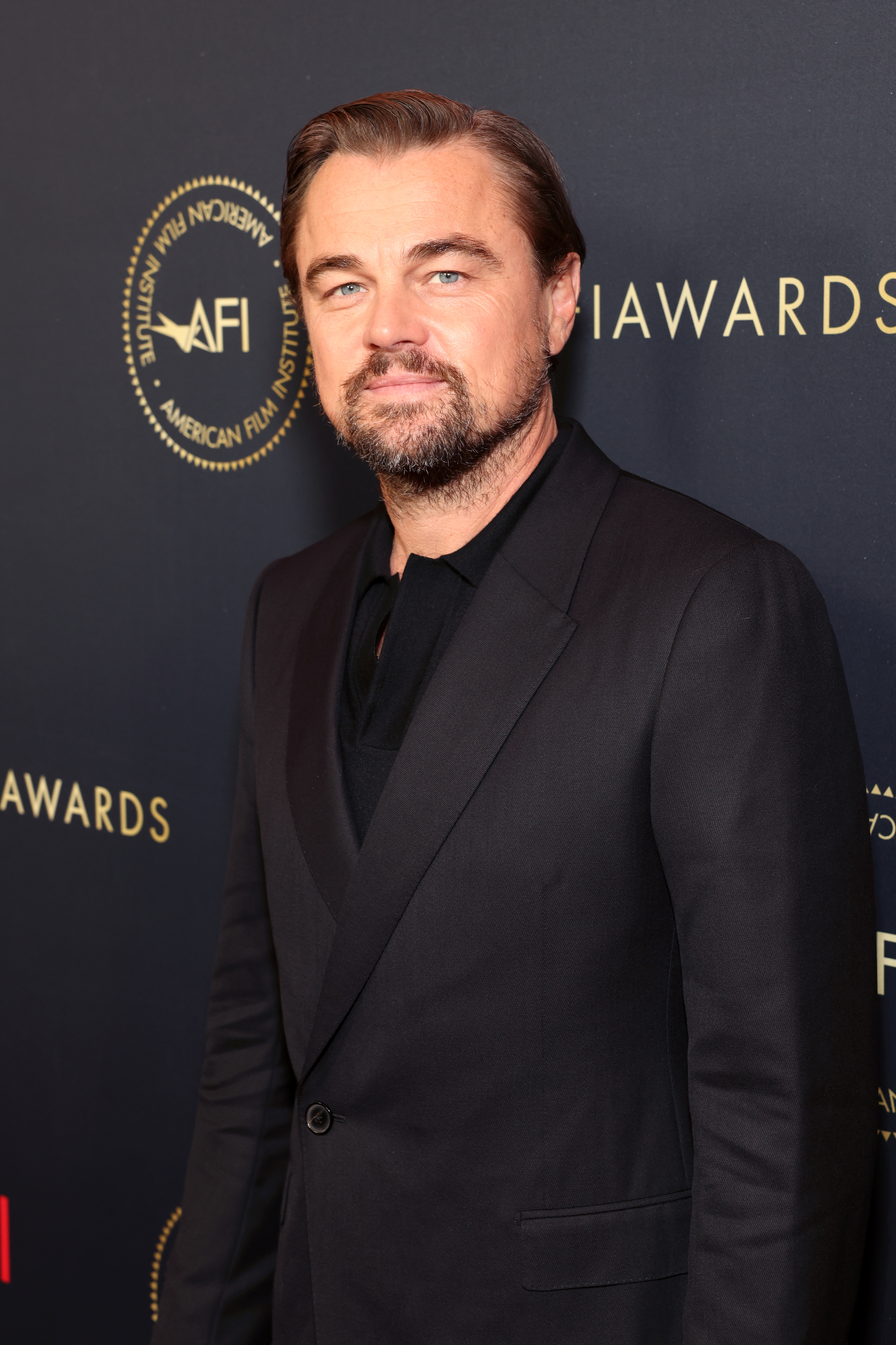 Leonardo DiCaprio at the AFI Awards, dressed in a classic black suit, posing in front of a branded backdrop