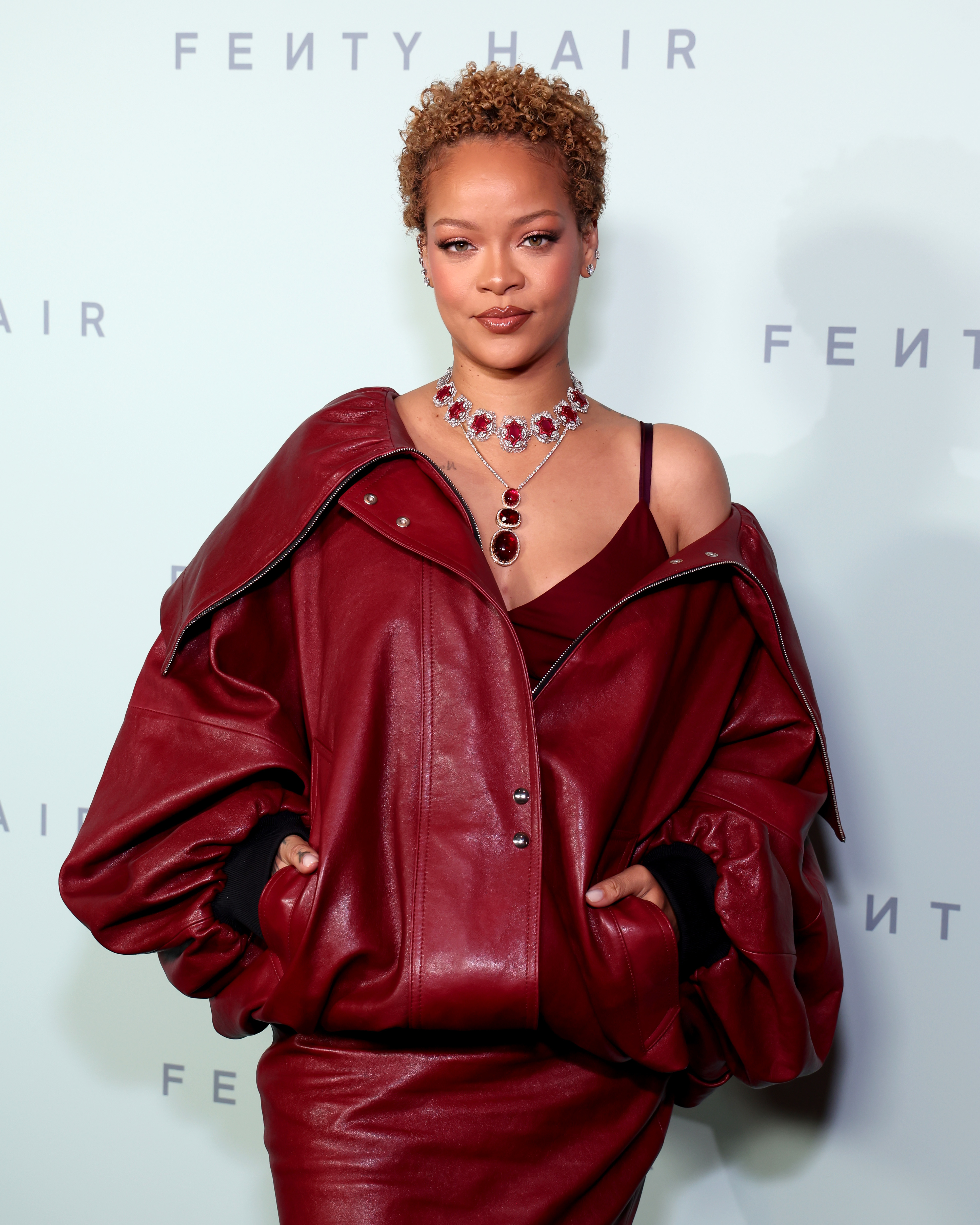 Rihanna poses on the red carpet wearing a large leather jacket, matching skirt, and a statement necklace at a Fenty Hair event