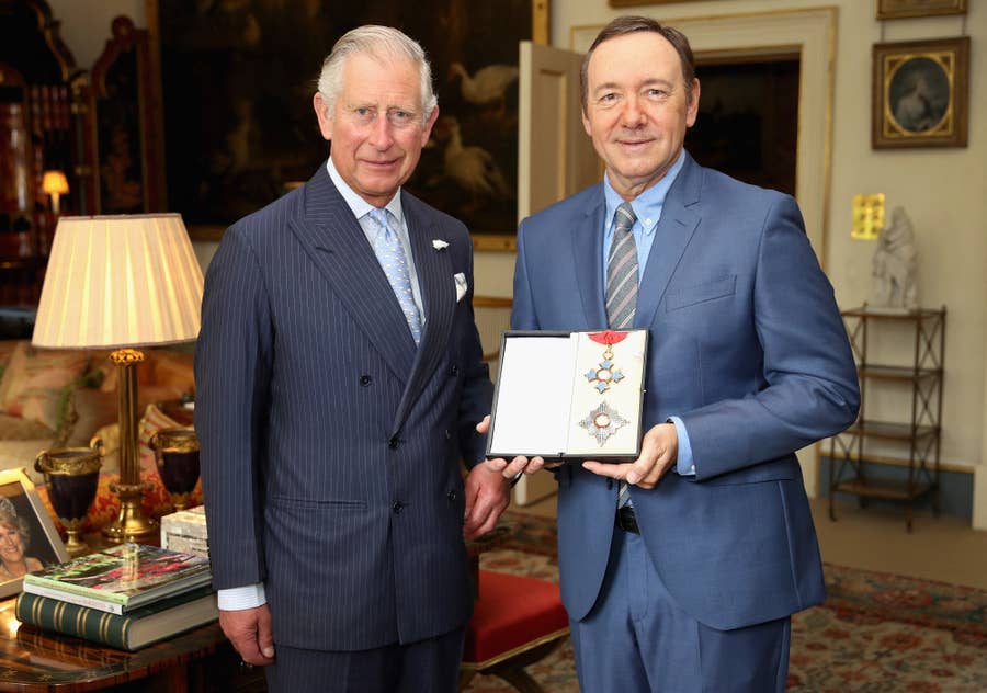Prince Charles and Kevin Spacey stand indoors, both wearing suits. Kevin Spacey holds an open award case displaying a medal