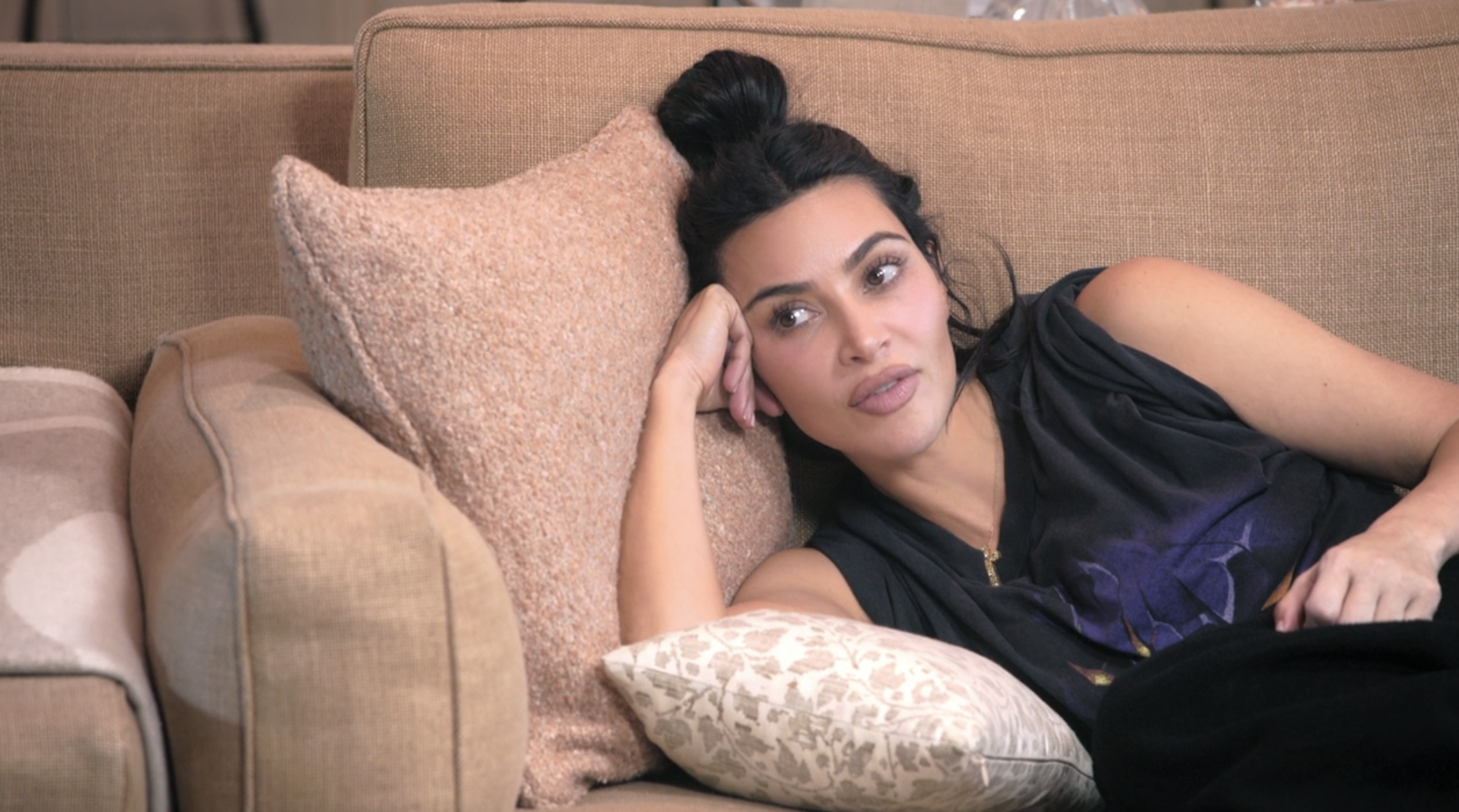 Kim Kardashian is lounging on a couch, resting her head on a pillow, looking off into the distance, wearing a sleeveless top