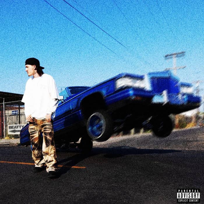 A person in a white shirt and baggy pants stands in front of a raised blue car on a street. The image includes a &quot;Parental Advisory&quot; label