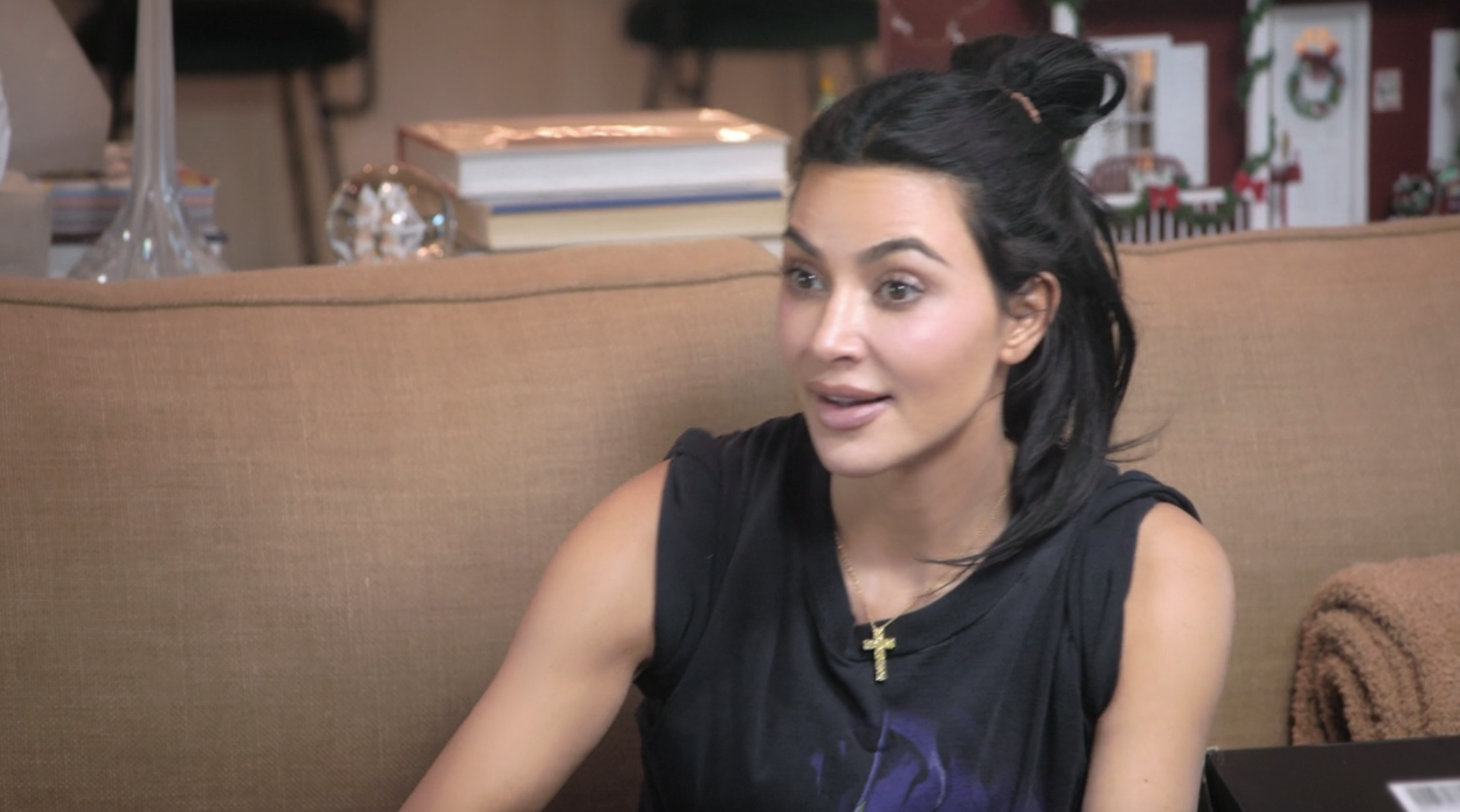 Kim Kardashian sits on a couch wearing a sleeveless dark top and a cross necklace, with a surprised expression