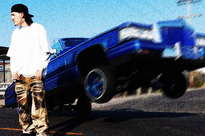 Person in streetwear and a backwards cap stands near a blue lowrider car lifting its front wheels in a street setting. Image is categorized as Music