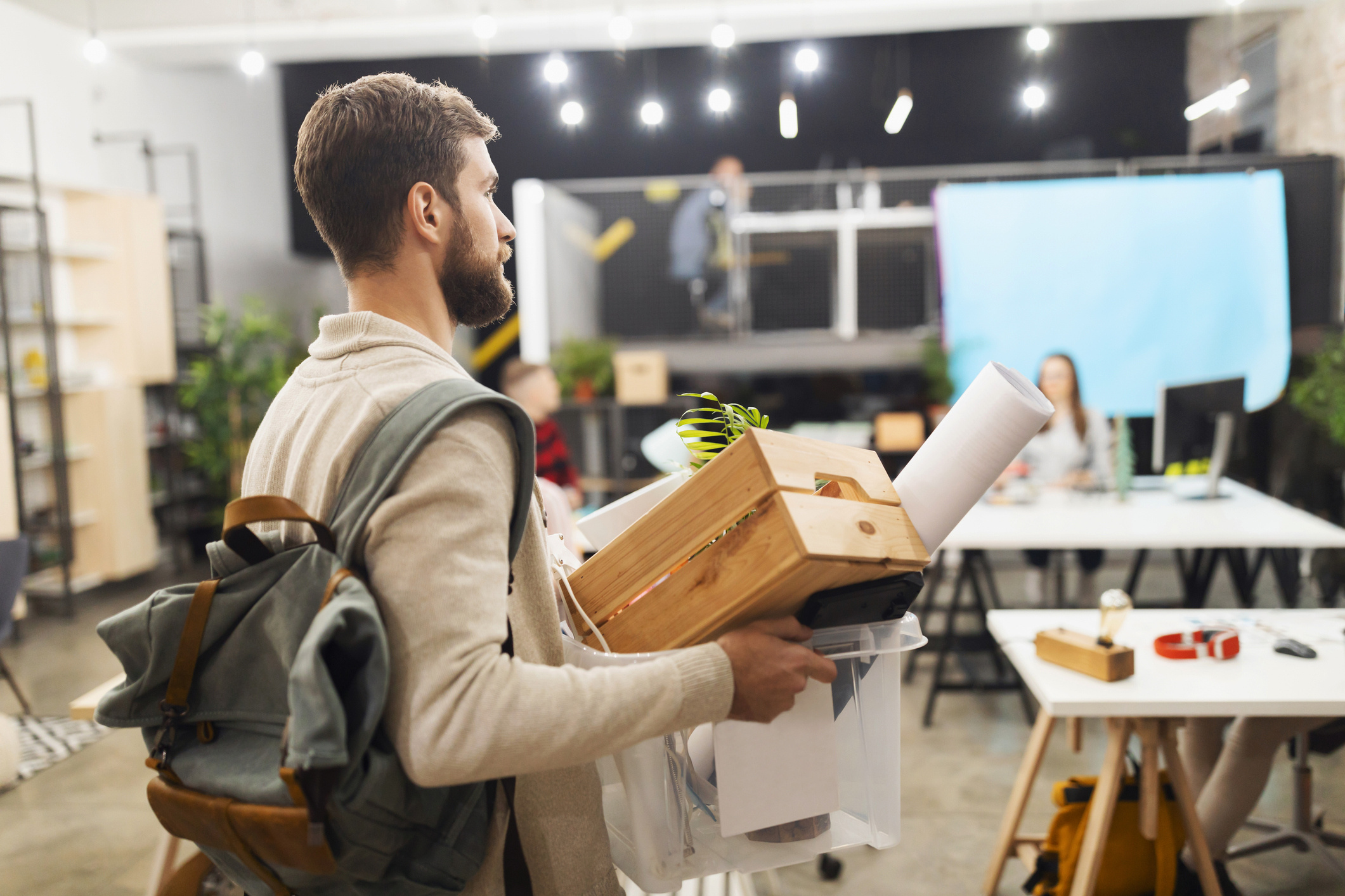A man carrying a backpack and a box filled with office supplies walks through an open-plan office space with blurred figures working in the background
