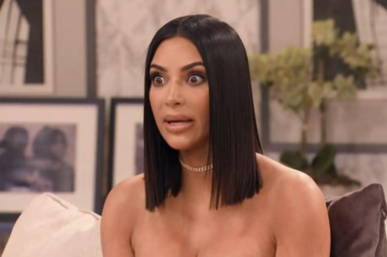 Kim Kardashian, with a surprised expression, wearing an off-shoulder outfit and a choker necklace, sitting indoors