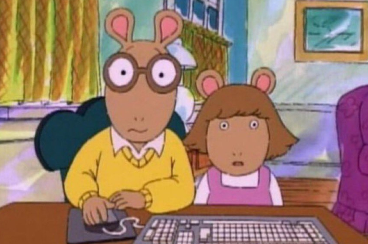 Arthur and D.W. from the animated series "Arthur" are at a computer, both looking surprised. Arthur wears glasses and a yellow sweater, and D.W. wears a pink dress