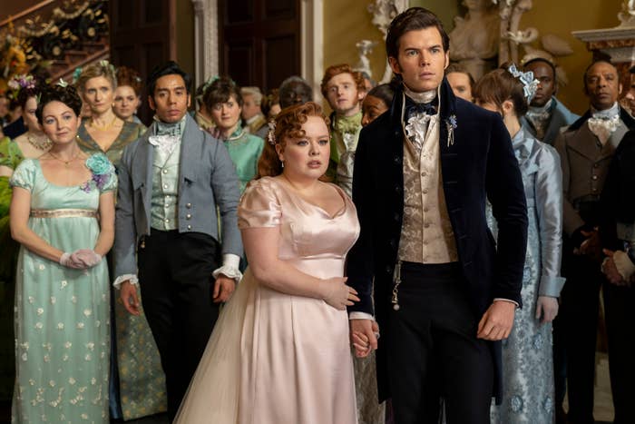 Group of actors in Regency-era costumes from Bridgerton, Simone Ashley, Nicola Coughlan, Luke Newton holding hands, among others, in an elegant hall
