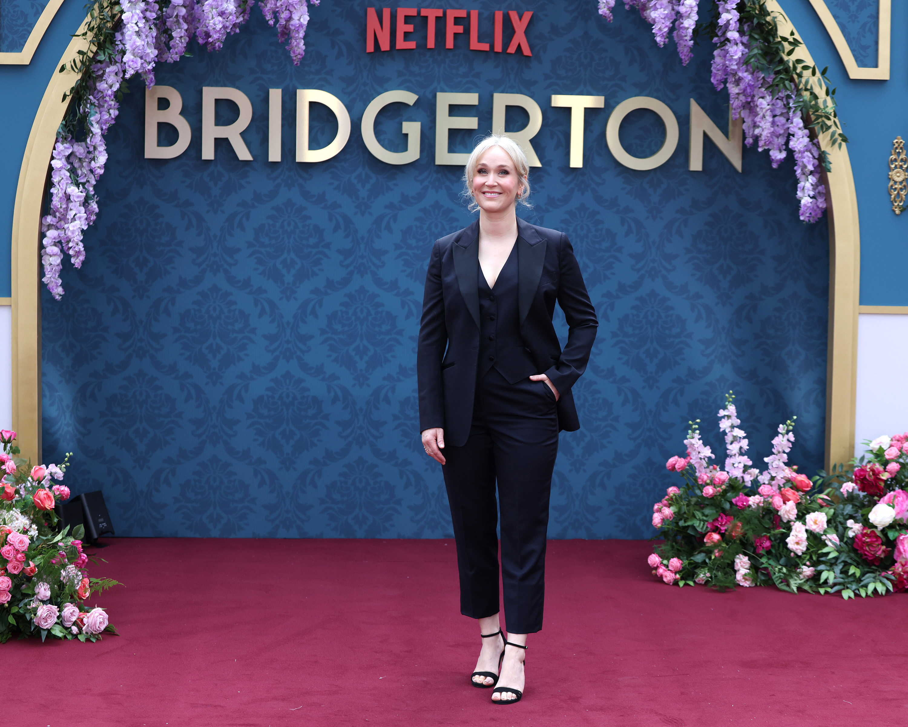 Jess Brownell poses on the red carpet in a black suit at the Bridgerton Netflix event, with floral decorations and the series title behind her