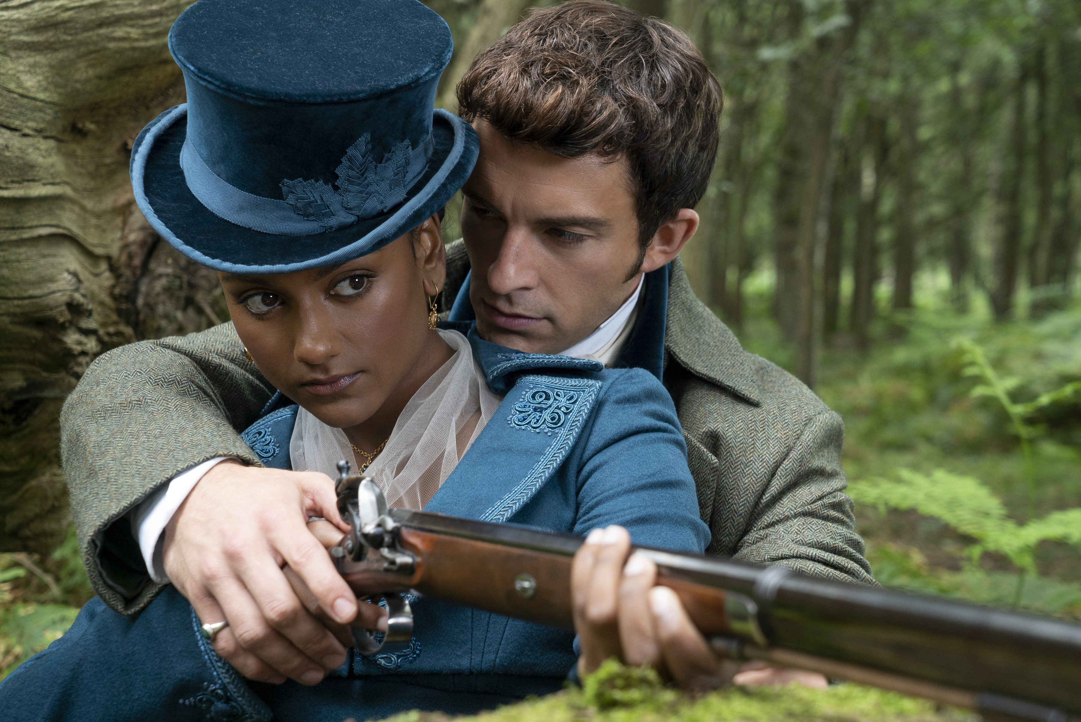 Simone Ashley and Jonathan Bailey, dressed in period costumes, sit in a forest as Simone's character aims a rifle with Jonathan's character assisting
