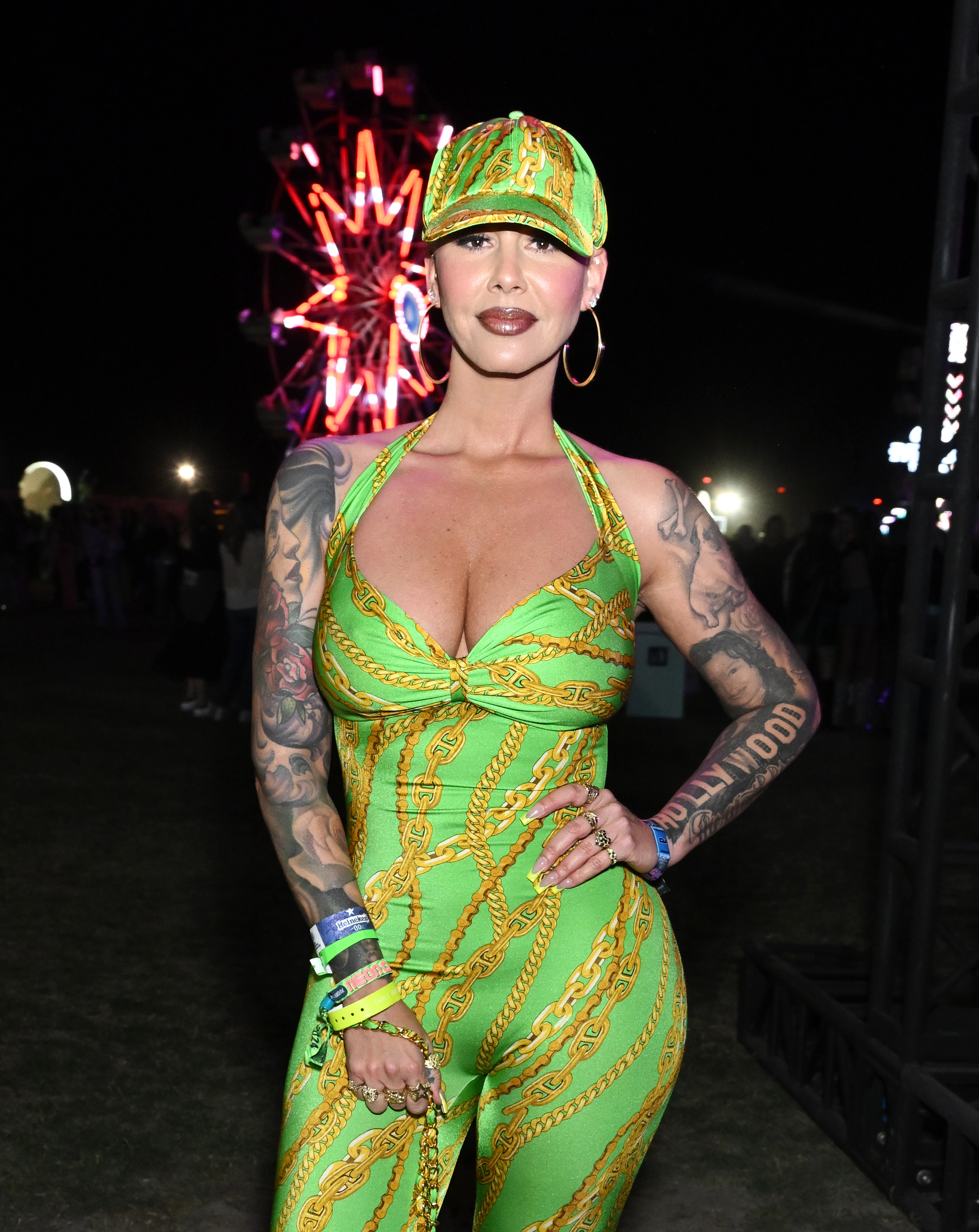 A woman in a patterned jumpsuit and matching cap poses outdoors. She has visible tattoos on her arms. A Ferris wheel is lit up in the background