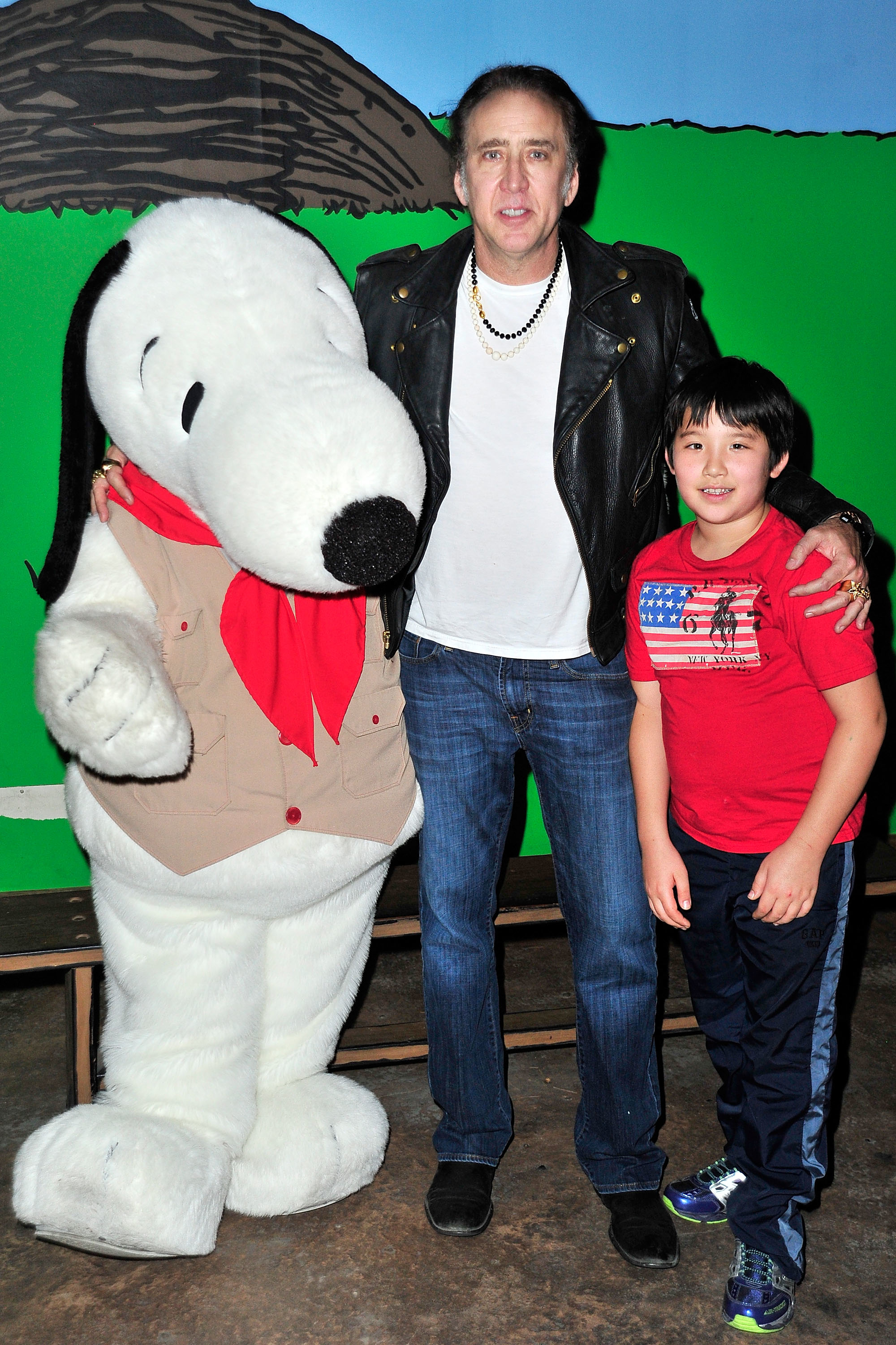Nicolas Cage posing with his son and a person in a Snoopy costume at an event. Nicolas Cage is wearing a black leather jacket and a white t-shirt