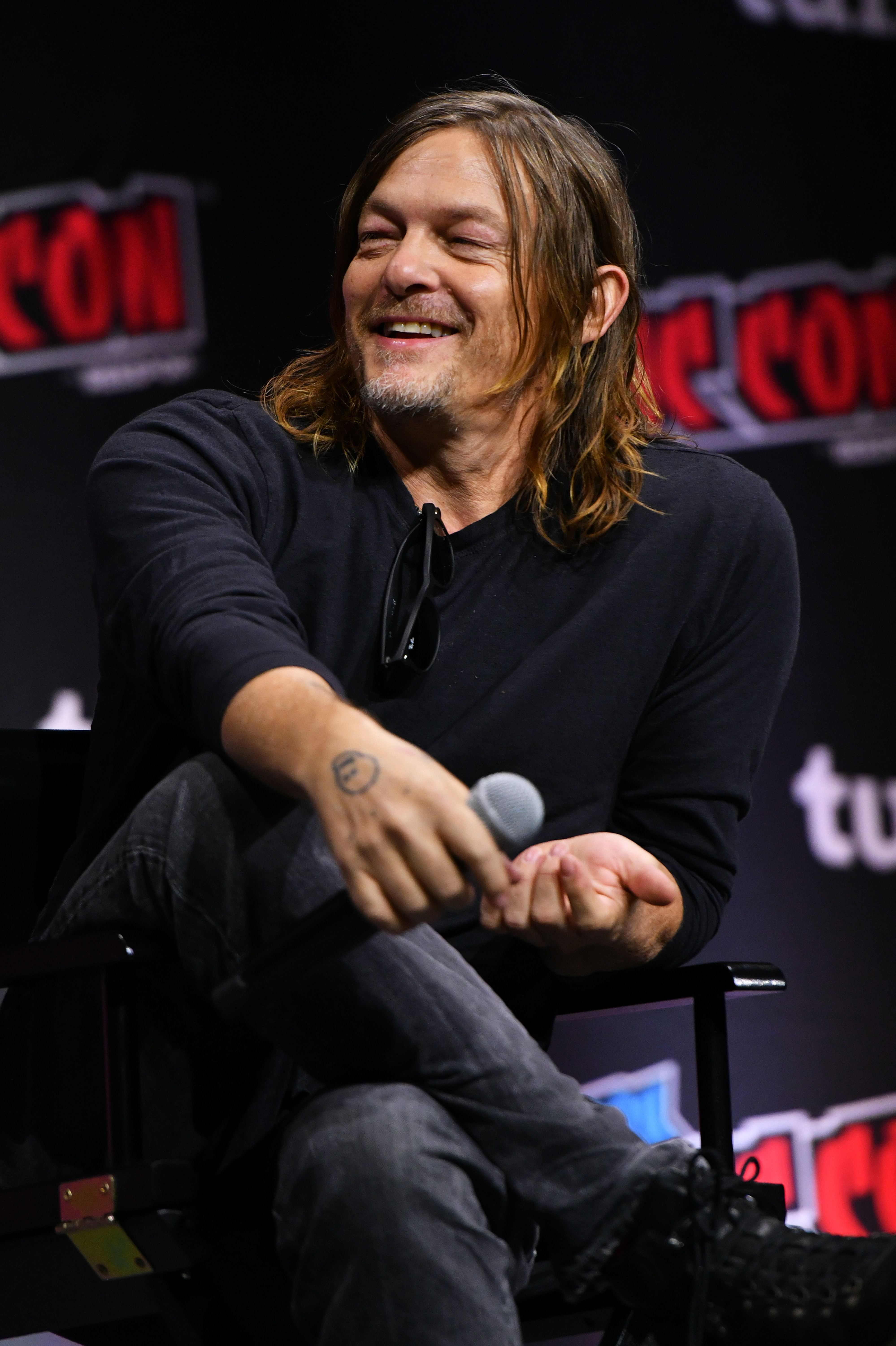 Norman Reedus sits on stage at a comic convention, holding a microphone and smiling, wearing a casual black long-sleeve shirt and grey jeans