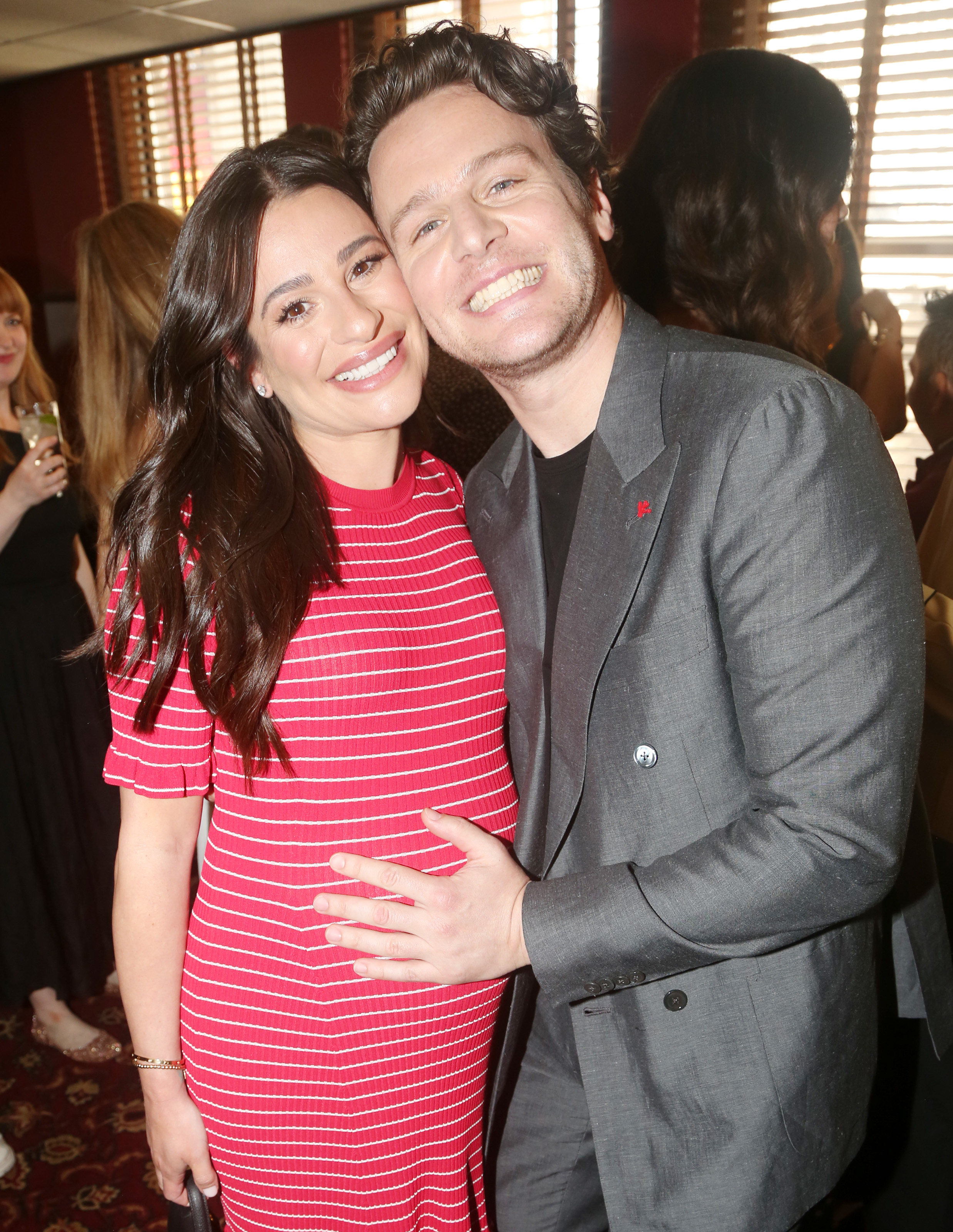 Lea Michele and Jonathan Groff pose together, smiling. She wears a striped dress, and he is in a gray suit with a black shirt. She is showing her baby bump