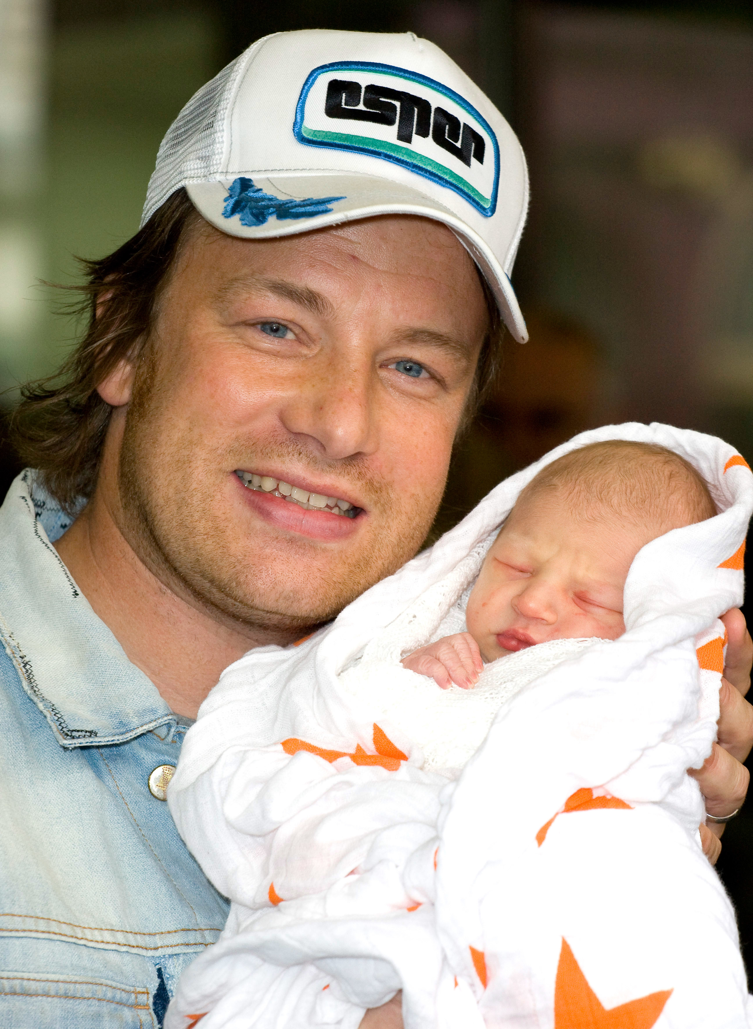 Jamie Oliver holding his newborn baby, wrapped in a white blanket with orange patterns, and smiling at the camera while wearing a casual cap and a denim jacket