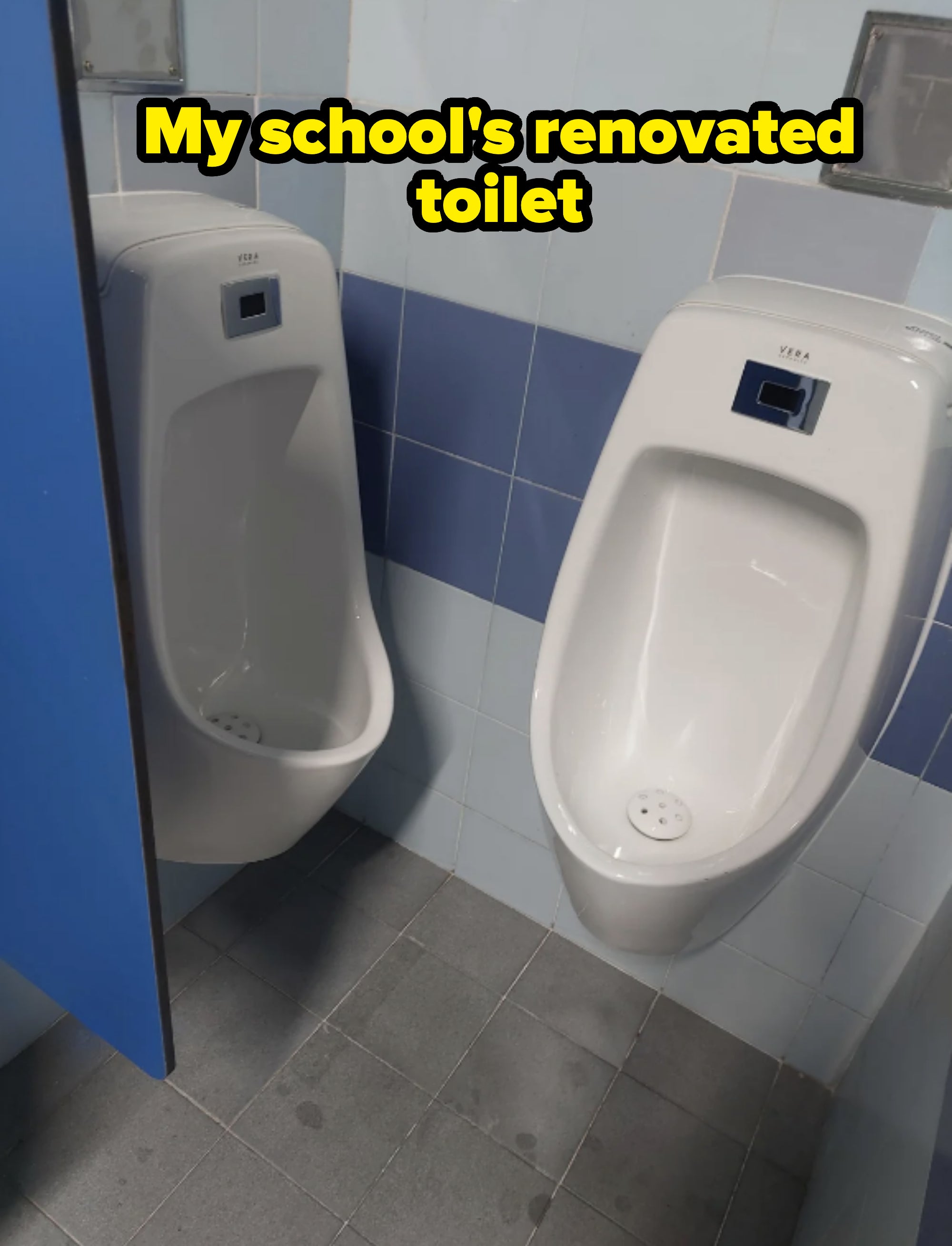 Two standard white urinals are installed side by side in a tiled public restroom. The restroom has light blue and white tiles