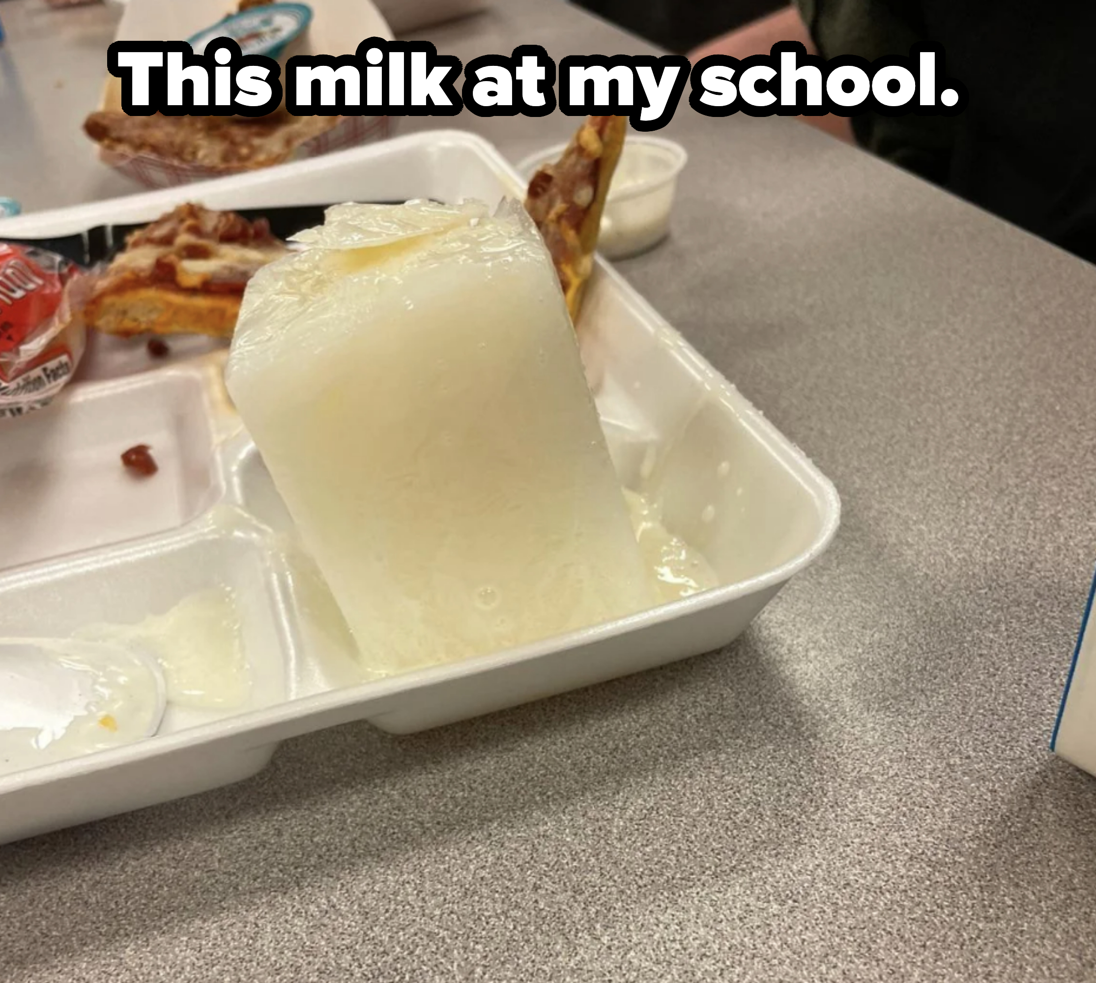A school lunch tray with a partially melted milk block, pizza slice with bacon, marinara sauce cup, and a container of dipping sauce