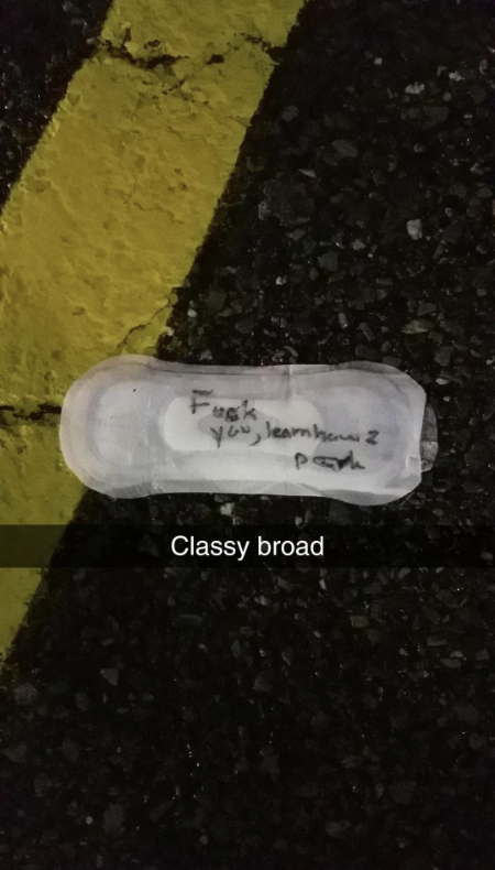 A sanitary pad with handwritten text reads, &quot;Fuck you ignorant peon.&quot; Below it, another text reads, &quot;Classy broad.&quot; The pad is on a road with yellow lines