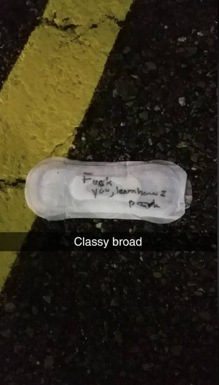 A sanitary pad with handwritten text reads, "Fuck you ignorant peon." Below it, another text reads, "Classy broad." The pad is on a road with yellow lines