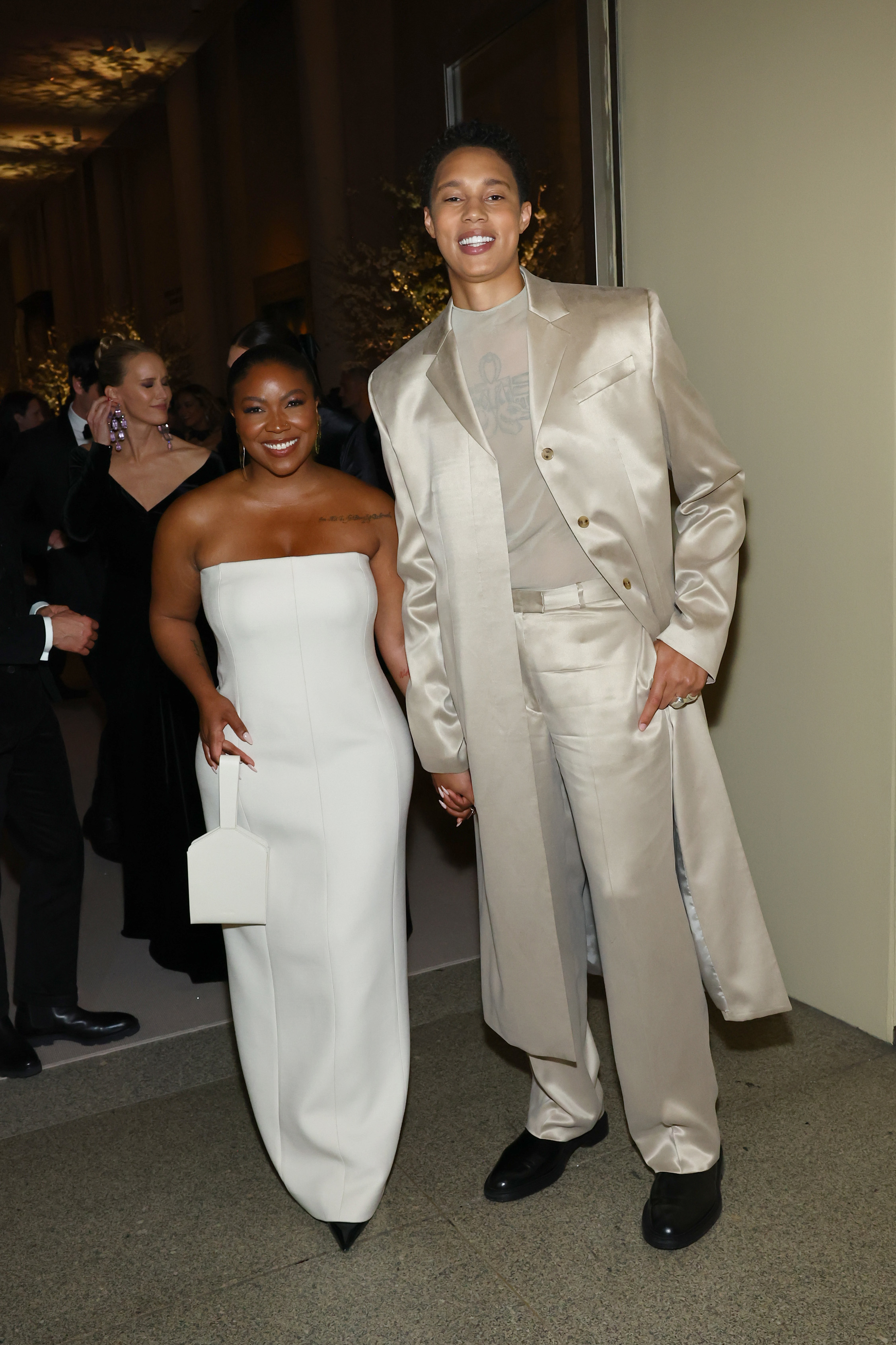 Brittney Griner and Cherelle Griner hold hands and smile at a red carpet event. Cherelle wears a strapless gown, Brittney wears a suit with a long coat
