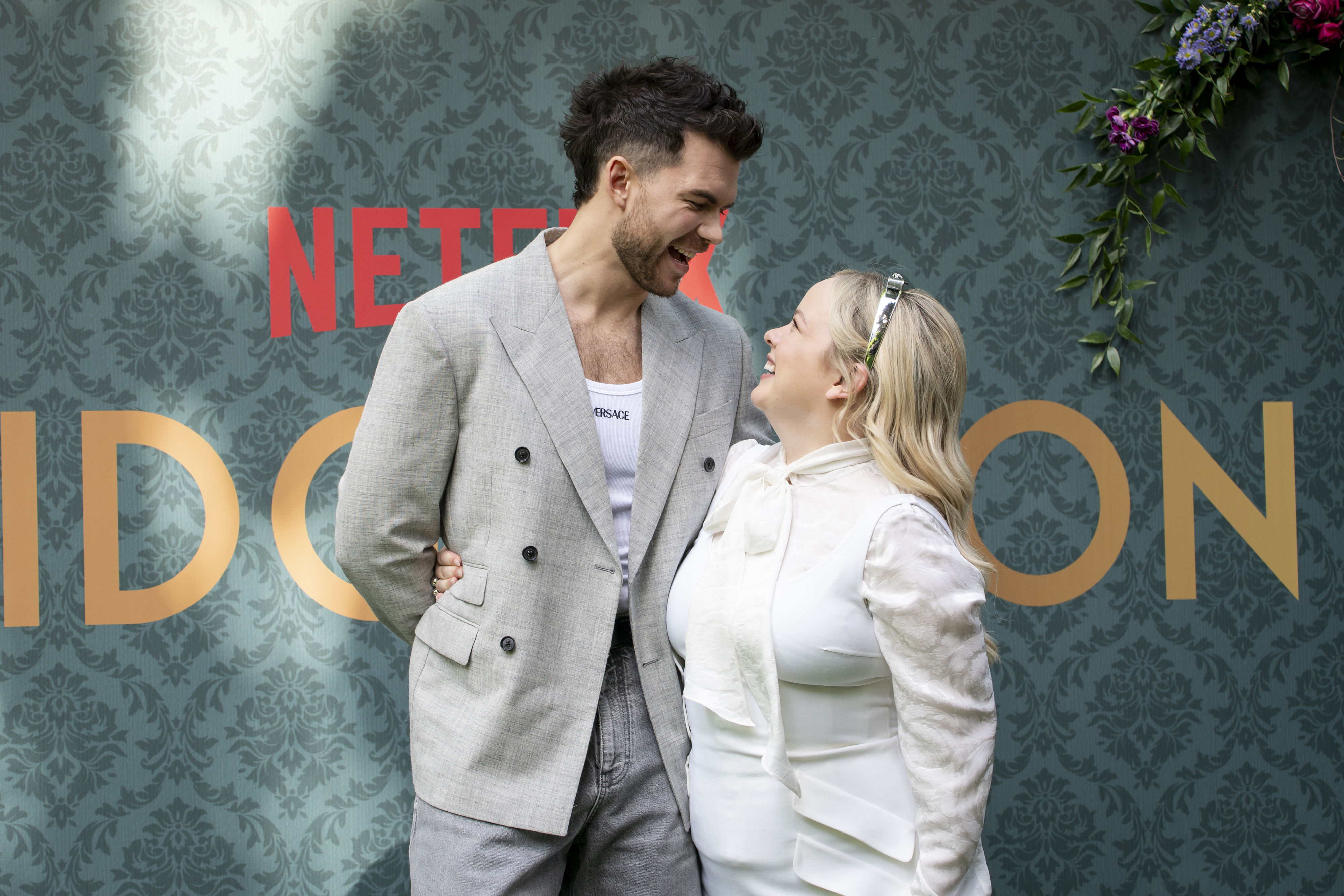 Simone Ashley and Jonathan Bailey smile at each other at a Netflix Bridgerton event. Simone is wearing a stylish gray blazer, and Jonathan is in a white blouse