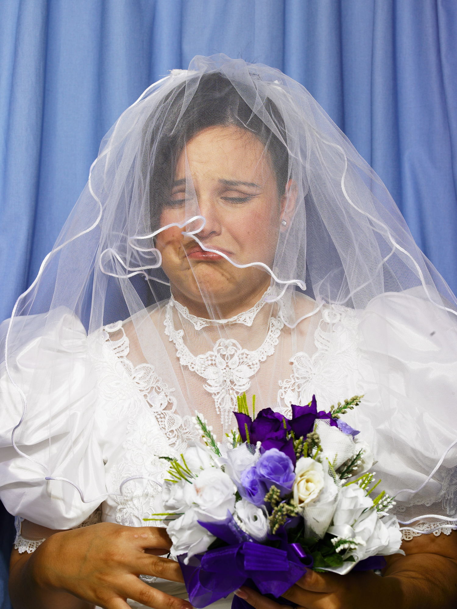 A bride in a wedding dress and veil is crying while holding a bouquet of white and purple flowers