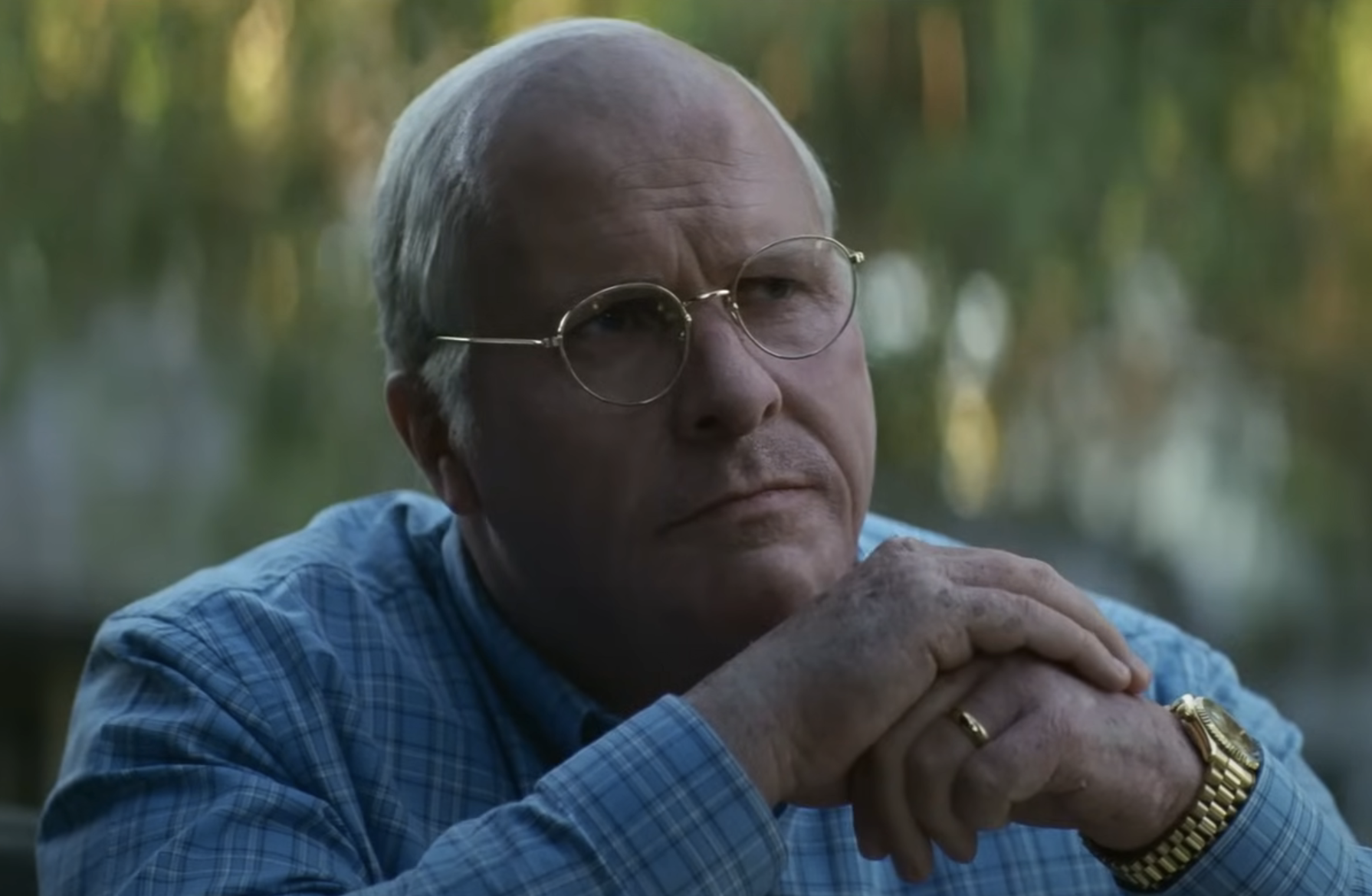 Christian Bale, in character, wearing glasses, a blue shirt, and a gold watch, rests his chin on his hands while staring thoughtfully