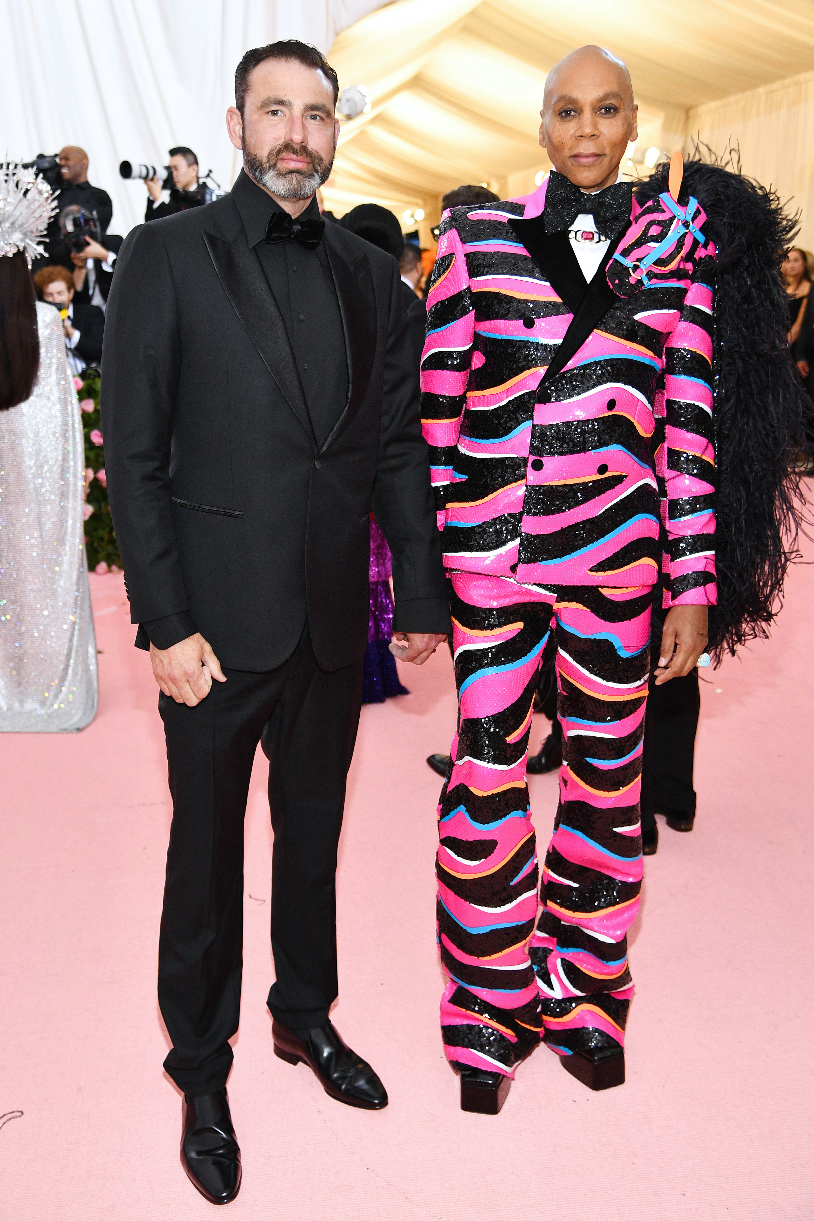 RuPaul and Georges LeBar stand together at a red carpet event. RuPaul wears a bold, patterned suit with feathered accents, while Georges LeBar wears a classic black tuxedo