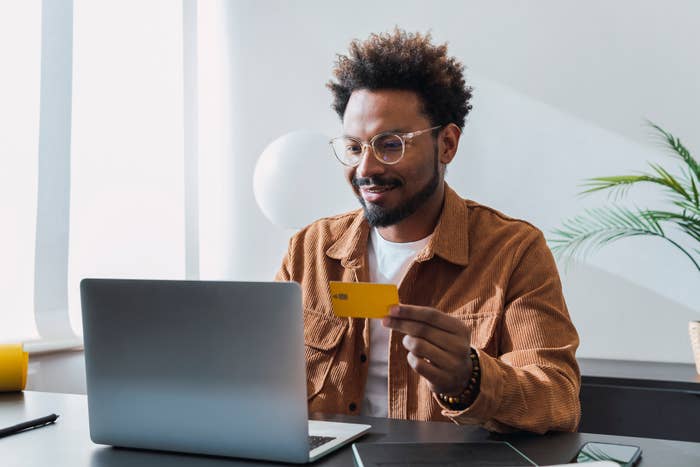 A man with glasses and a beard, wearing a casual jacket, sits at a desk using a laptop while holding a credit card