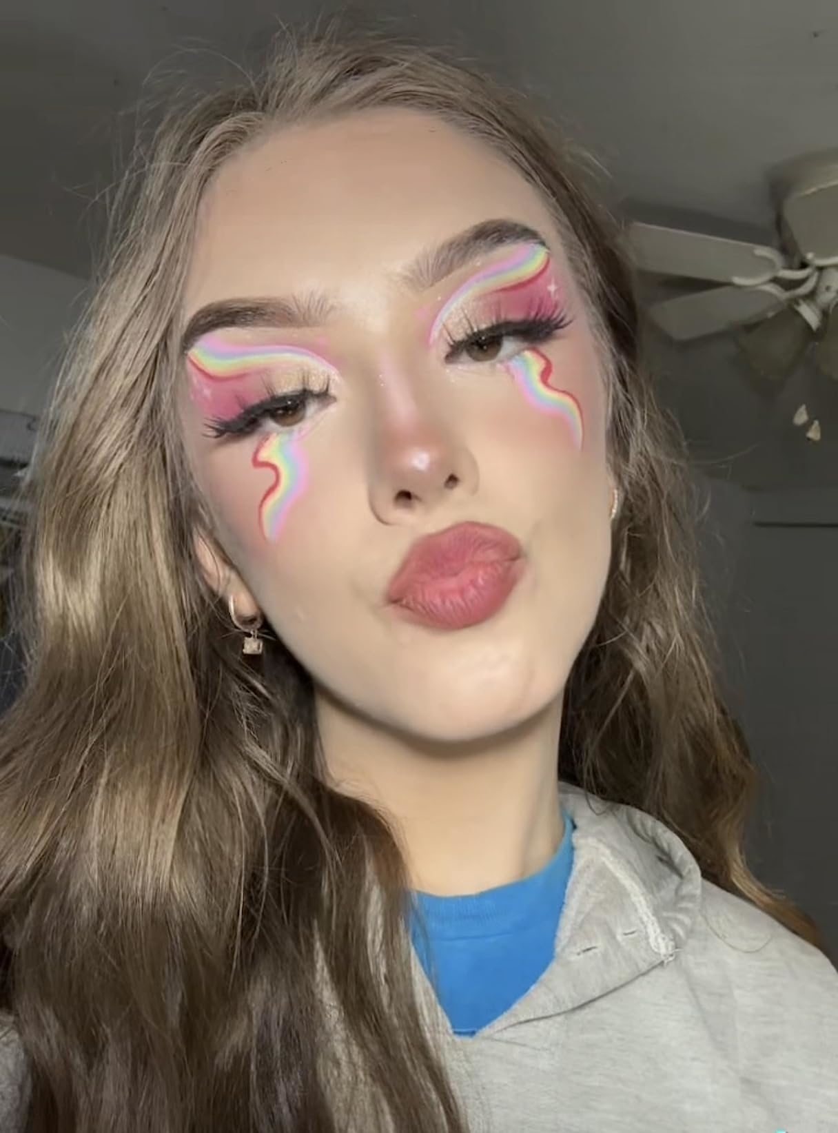 A reviewer with long hair wearing a hoodie, bold eye makeup with pink eyeshadow, and rainbow designs extending from their eyes