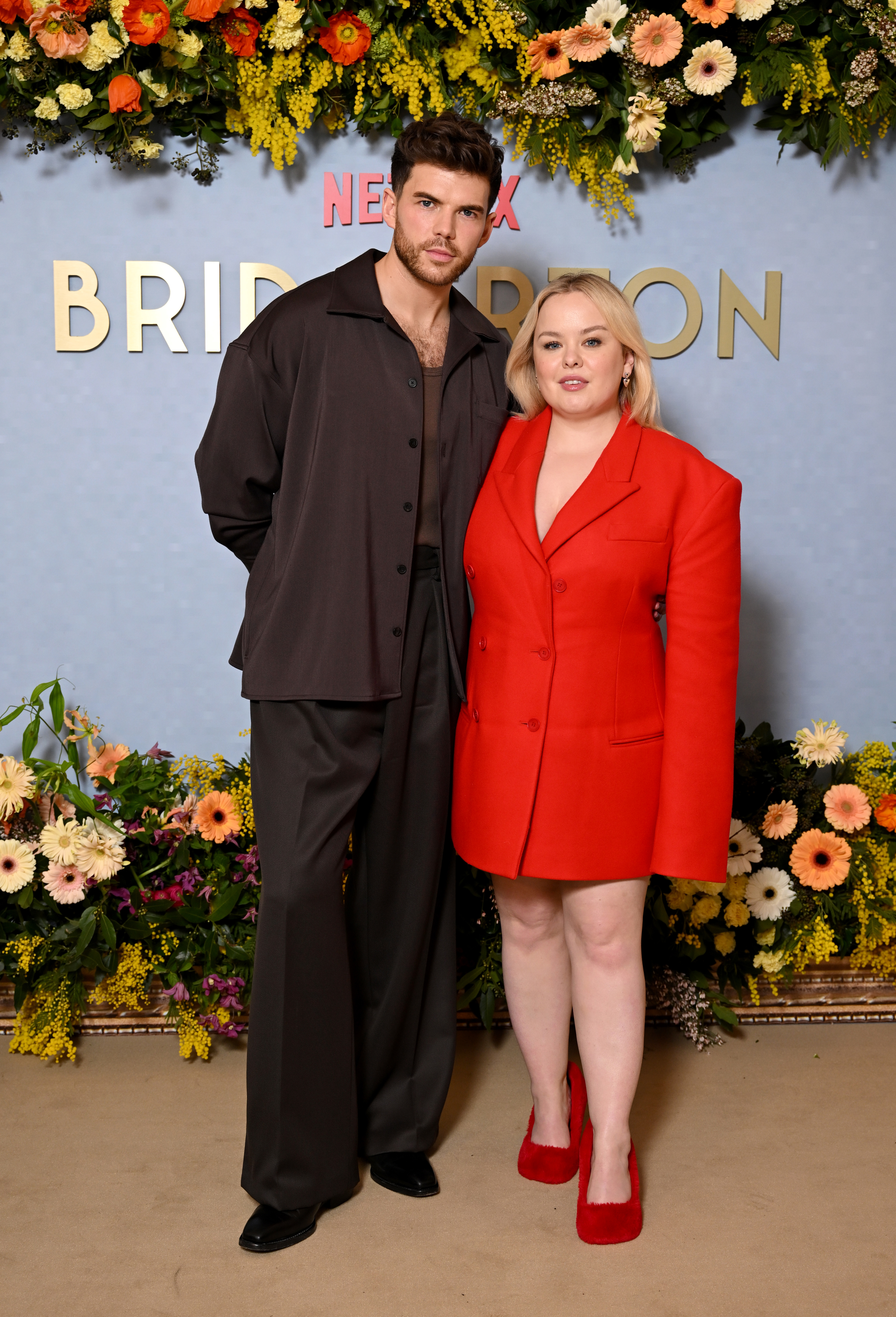 Nicola Coughlan and Luke Newton posed together at a Bridgerton event, standing in front of a floral backdrop. Nicola wears an oversized red blazer, while Luke is in a dark suit