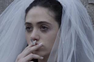 Emmy Rossum in a bridal veil and dress, smoking a cigarette with a pensive expression. 
