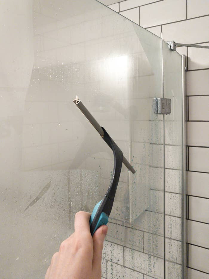 Hand using a squeegee to clean water-streaked glass shower door in a tiled bathroom