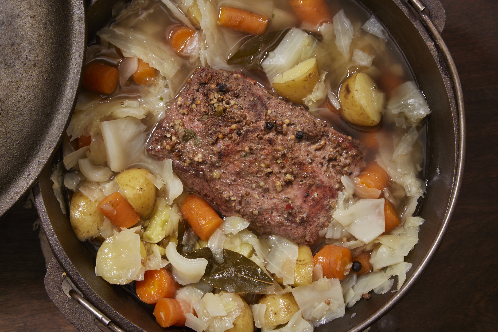 A close-up of a pot containing a cooked meal with a piece of seasoned meat, potatoes, carrots, cabbage, and various herbs