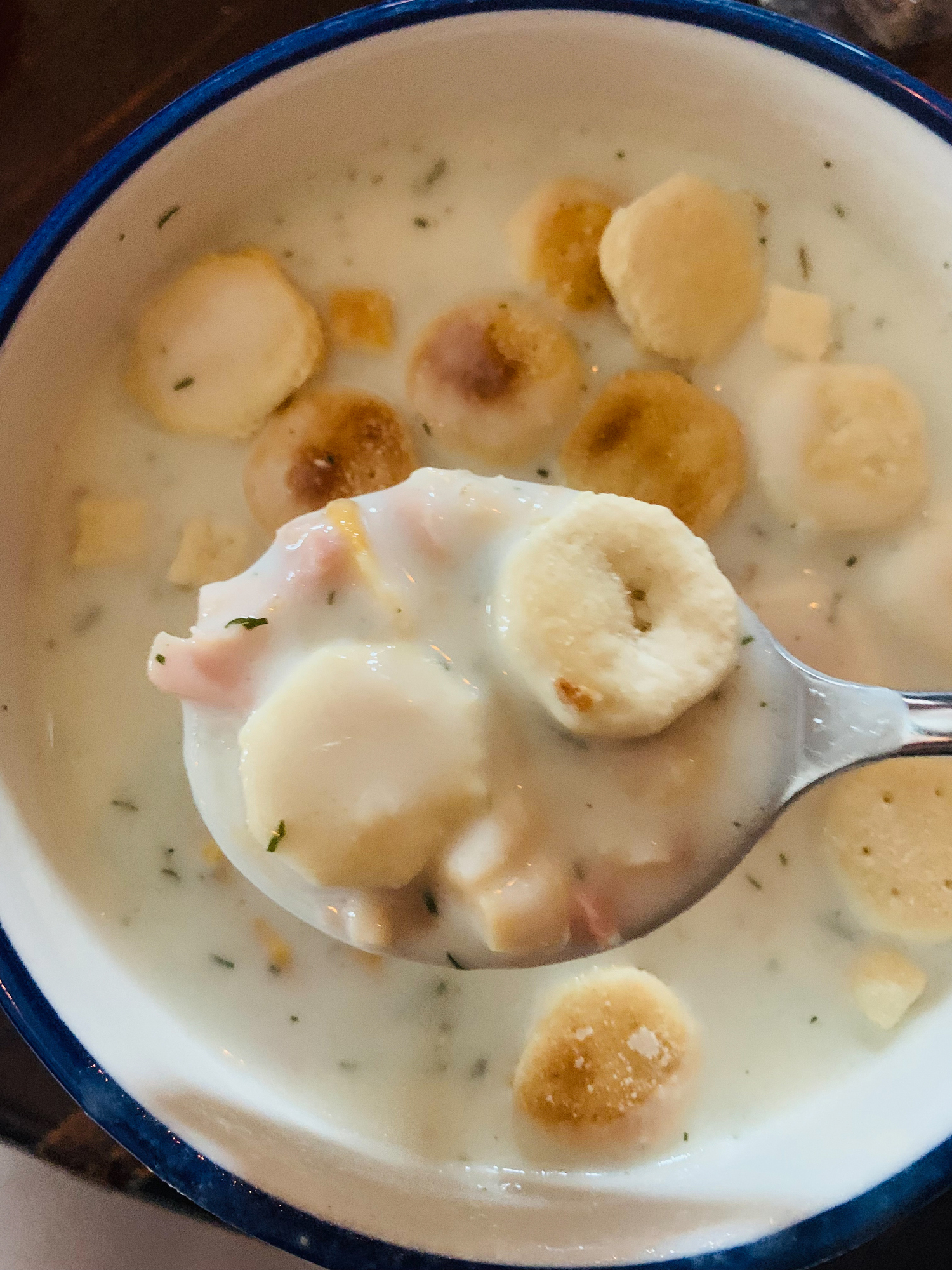 A bowl of creamy white soup with round crackers and bits of meat, with a spoon lifting some soup and crackers from the bowl