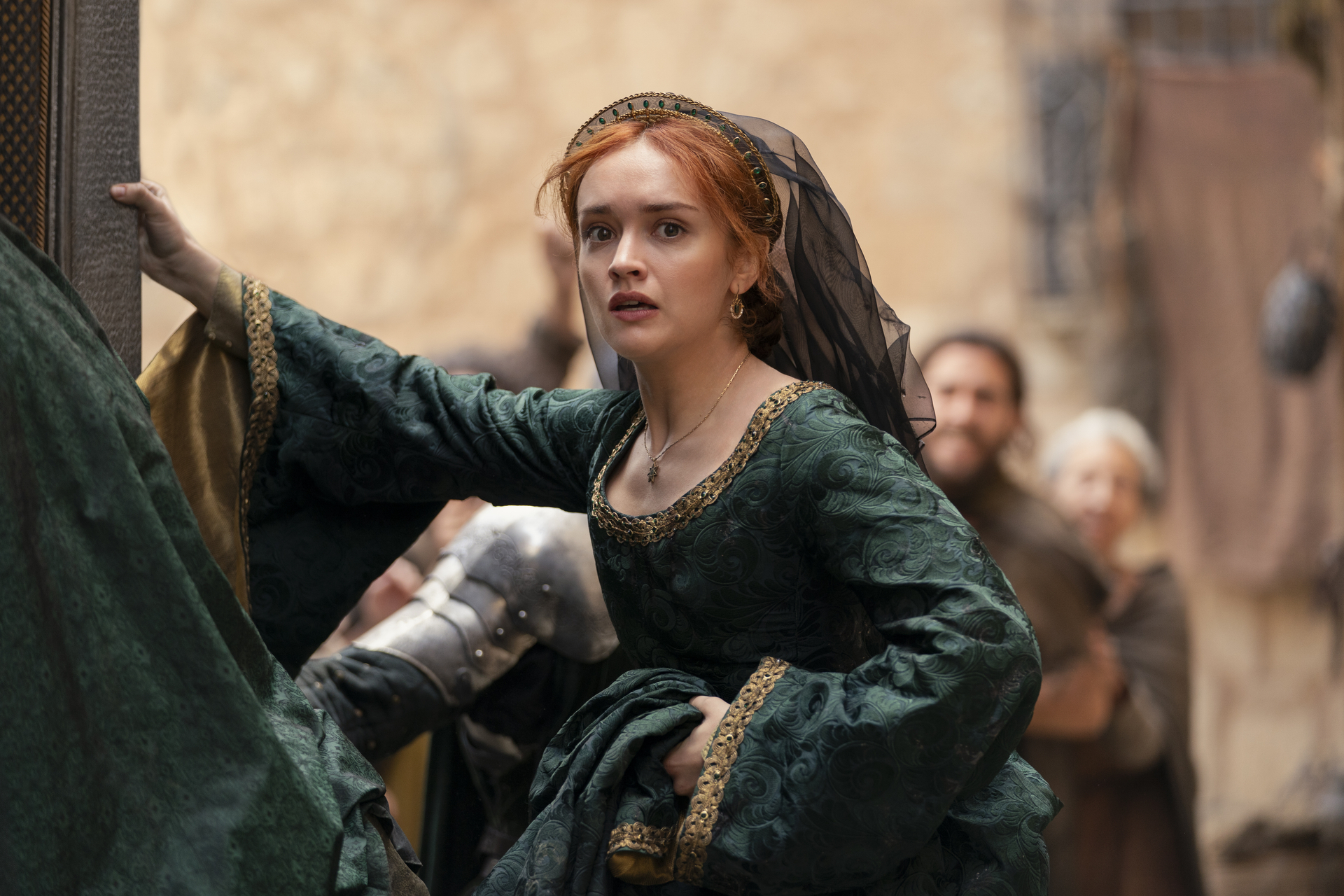 Olivia Cooke as Alicent Hightower is shown in a medieval-style green dress with gold embroidery, looking concerned in a historical setting with blurred figures in the background