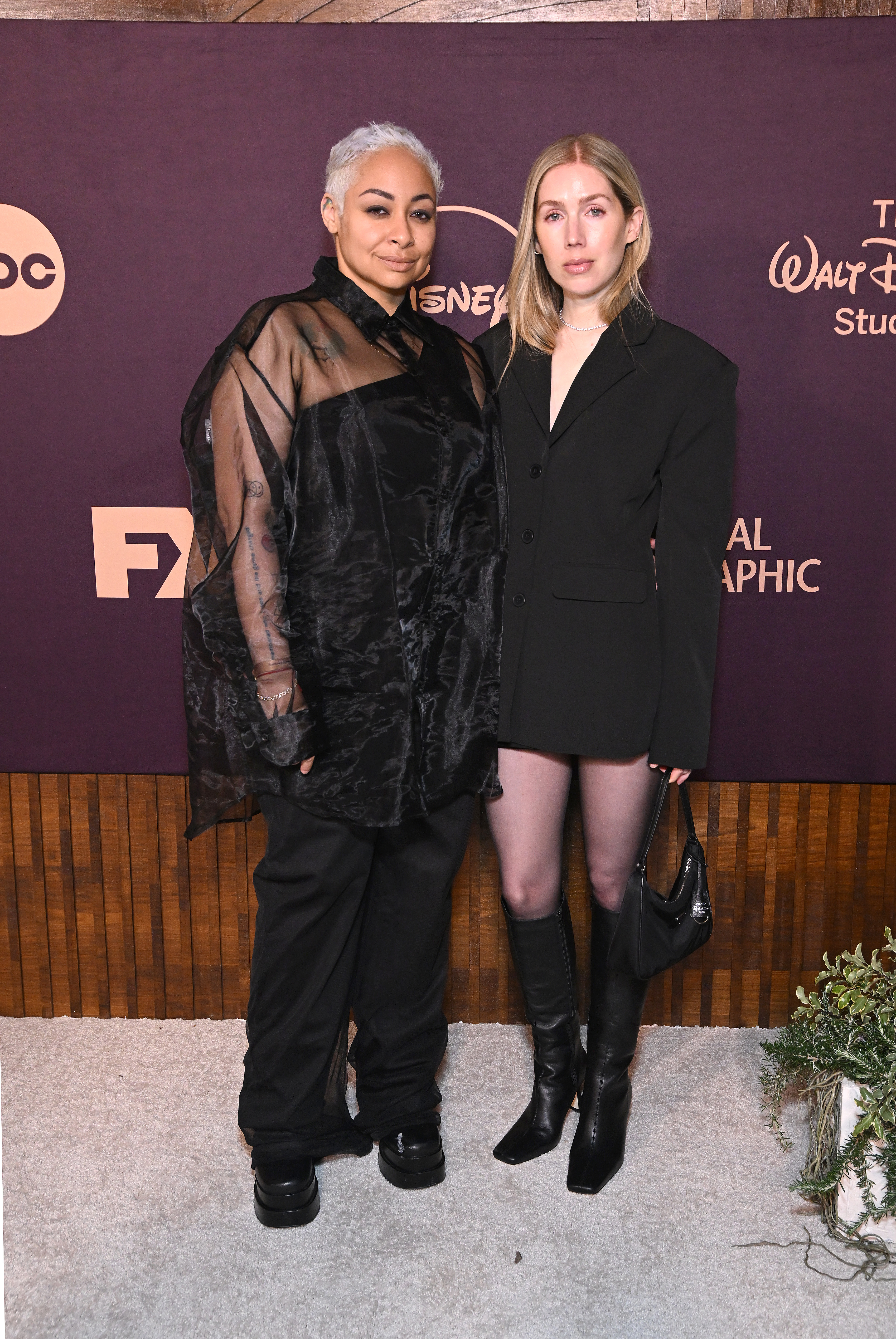 Raven-Symoné in a sheer black outfit and Miranda Maday in a black blazer and skirt, posing together at a Disney event