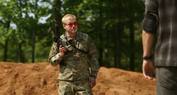Jesse, in military camouflage, holds a rifle, standing and talking to another person