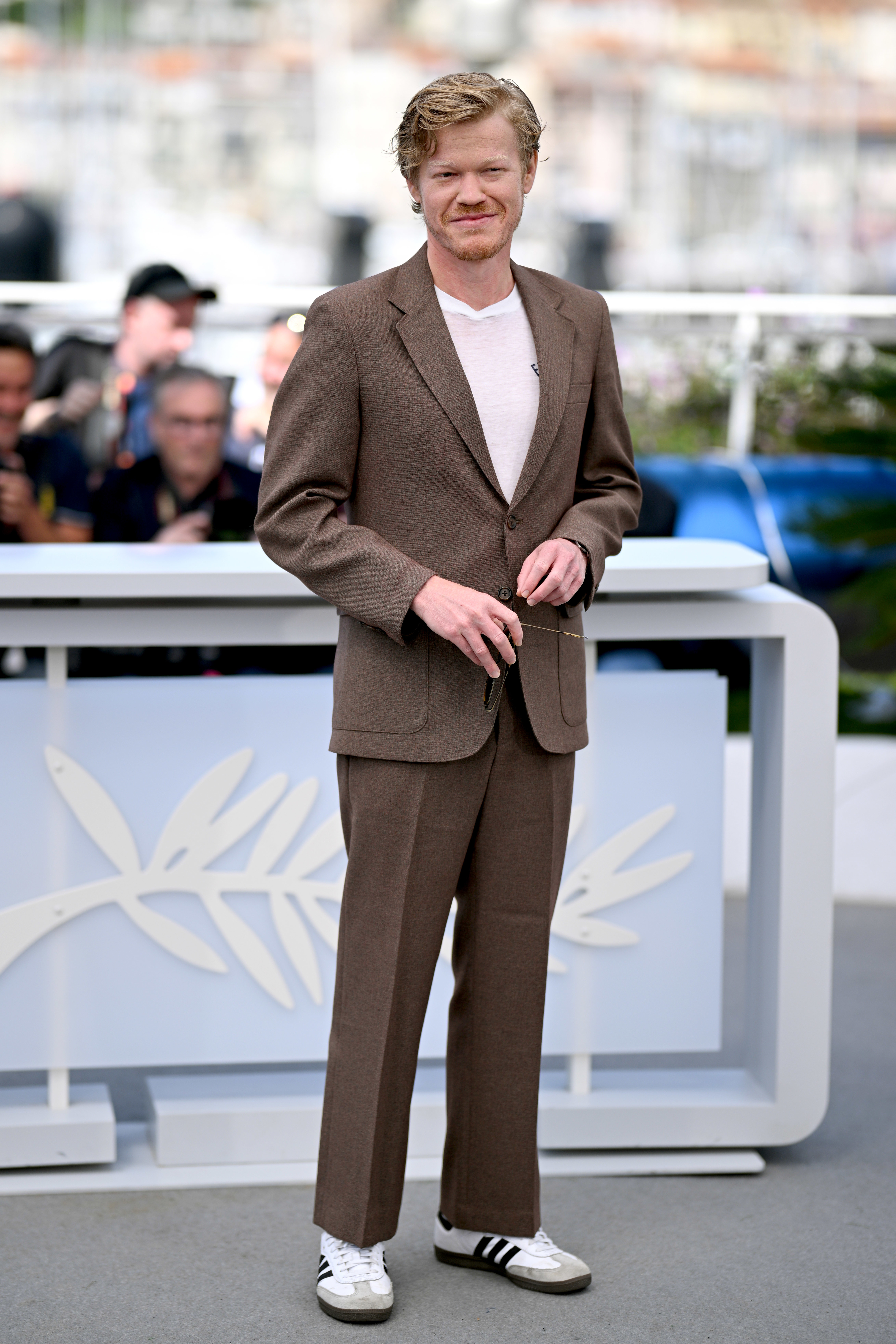 Jesse Plemons poses at an event, wearing a suit with a shirt and sneakers, standing in front of a backdrop featuring the Cannes Film Festival logo