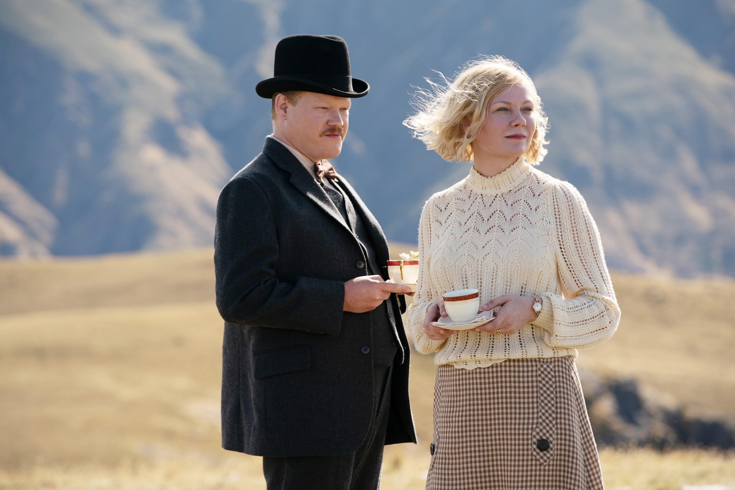 Jesse Plemons and Kirsten Dunst stand outdoors in period attire, holding teacups; mountains are visible in the background