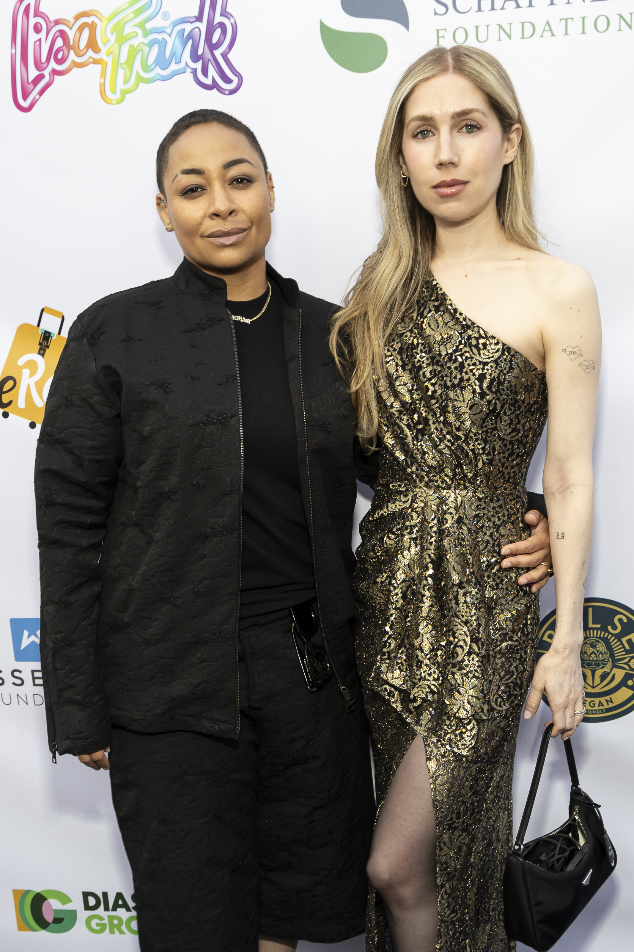 Raven-Symoné in a black outfit and Miranda Maday in a one-shoulder gold dress pose on the red carpet at an event