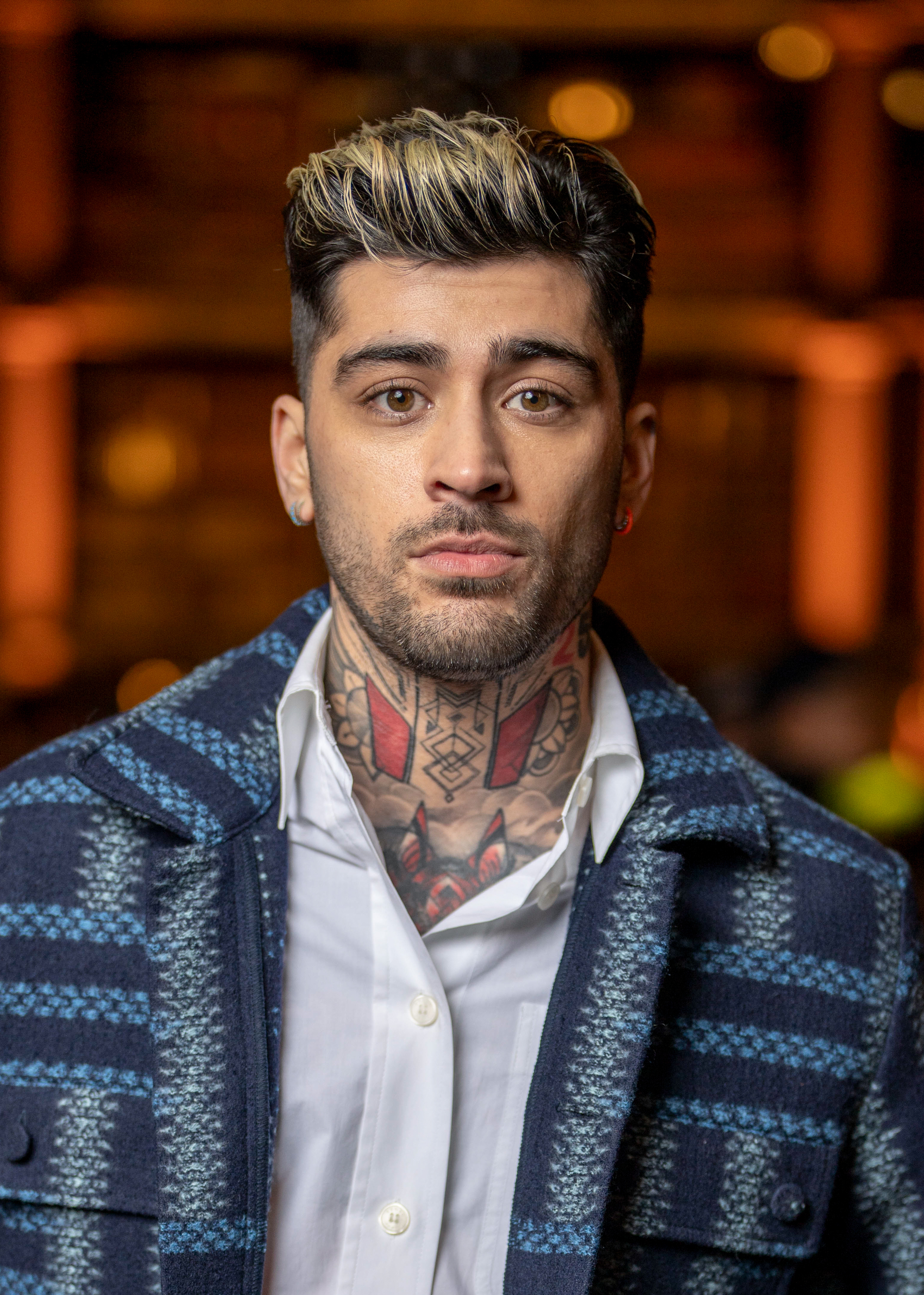 Zayn Malik poses for a photo wearing a white shirt under a plaid jacket, showcasing his neck tattoos