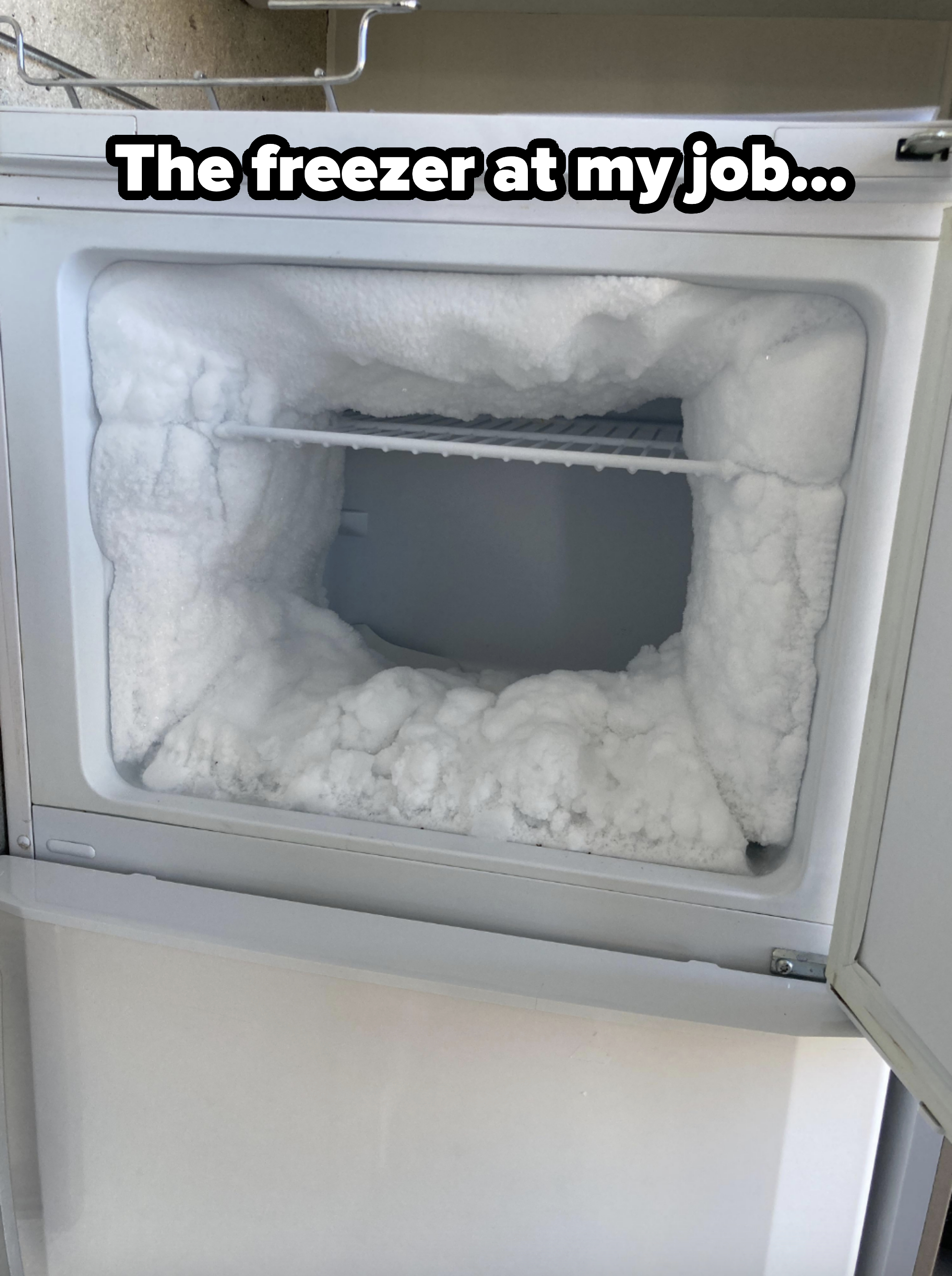 An open freezer with significant ice buildup on all interior surfaces, partially obstructing the shelf inside